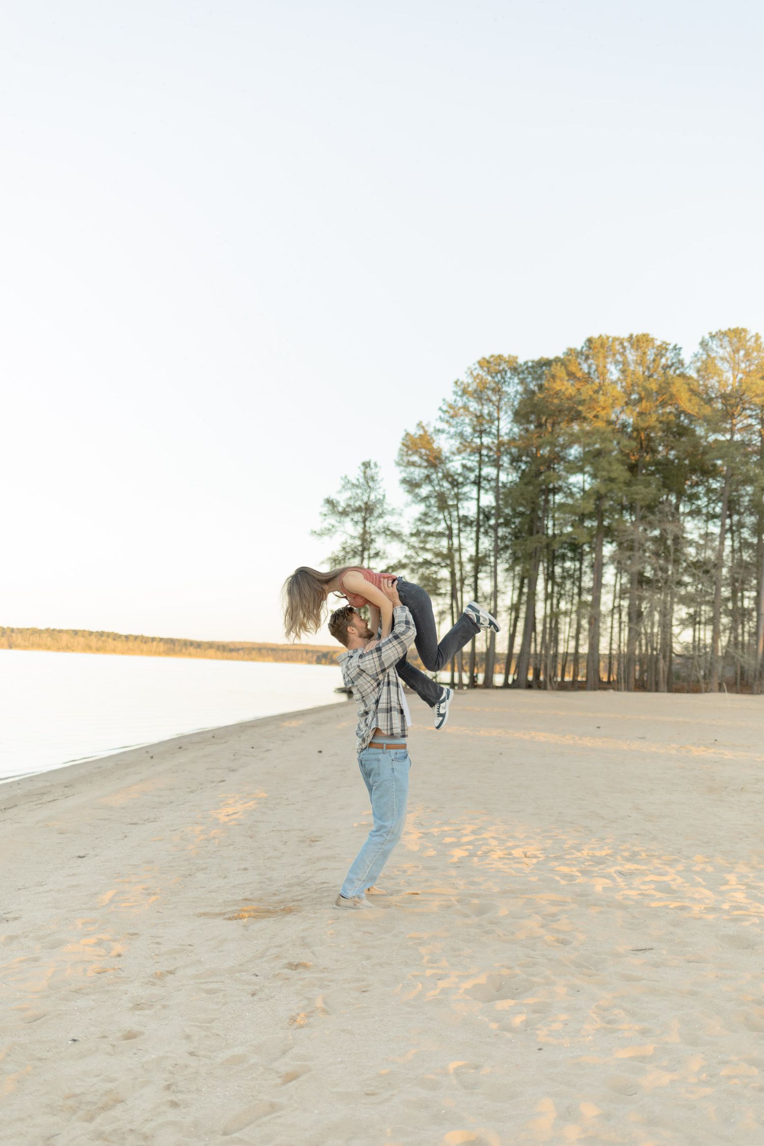 A playful lift on a sandy beach with pine trees and water in the background during golden hour during their couples lifestyle photoshoot.