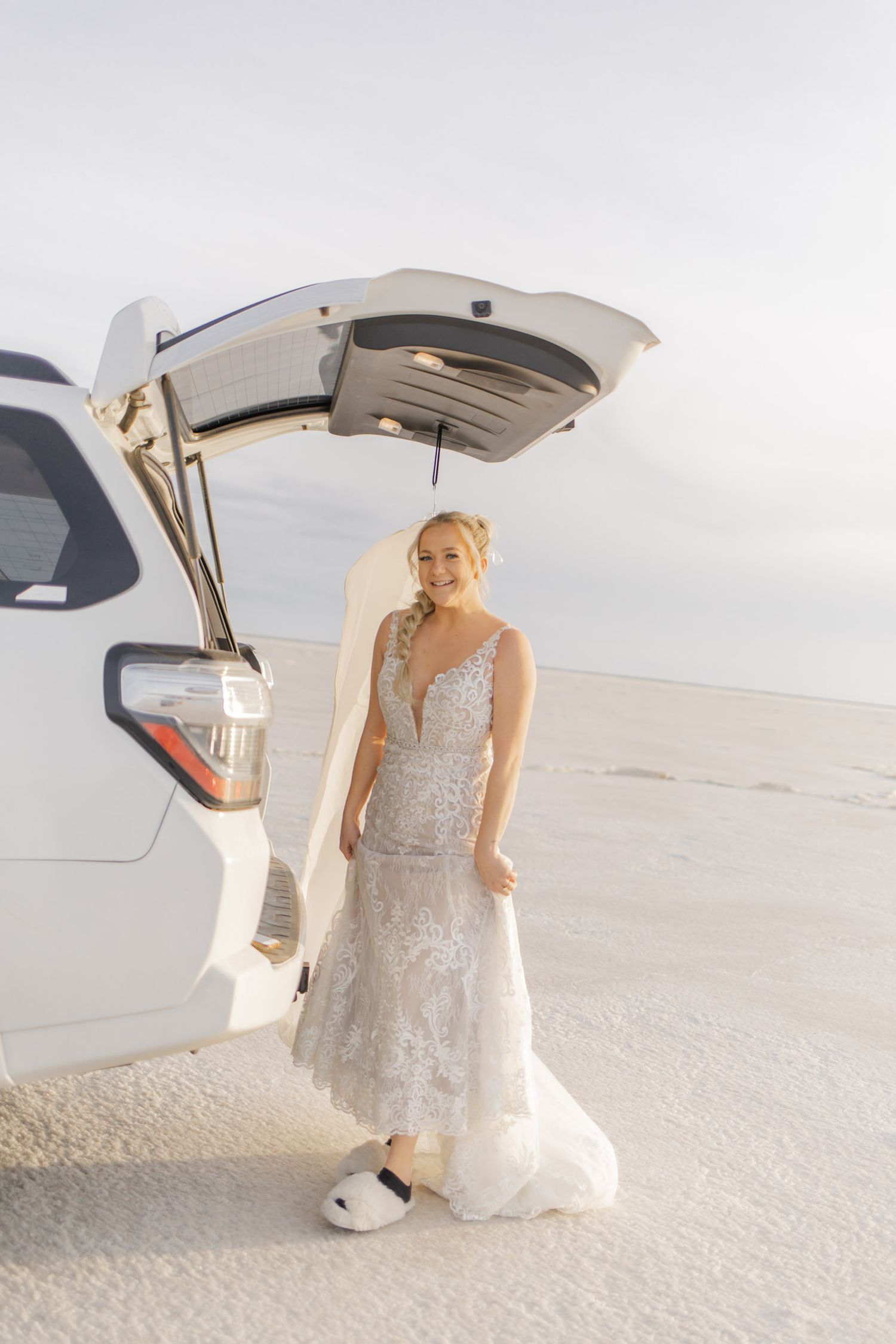 Person in an elegant lace wedding gown standing near the open trunk of a white car on salt flats.