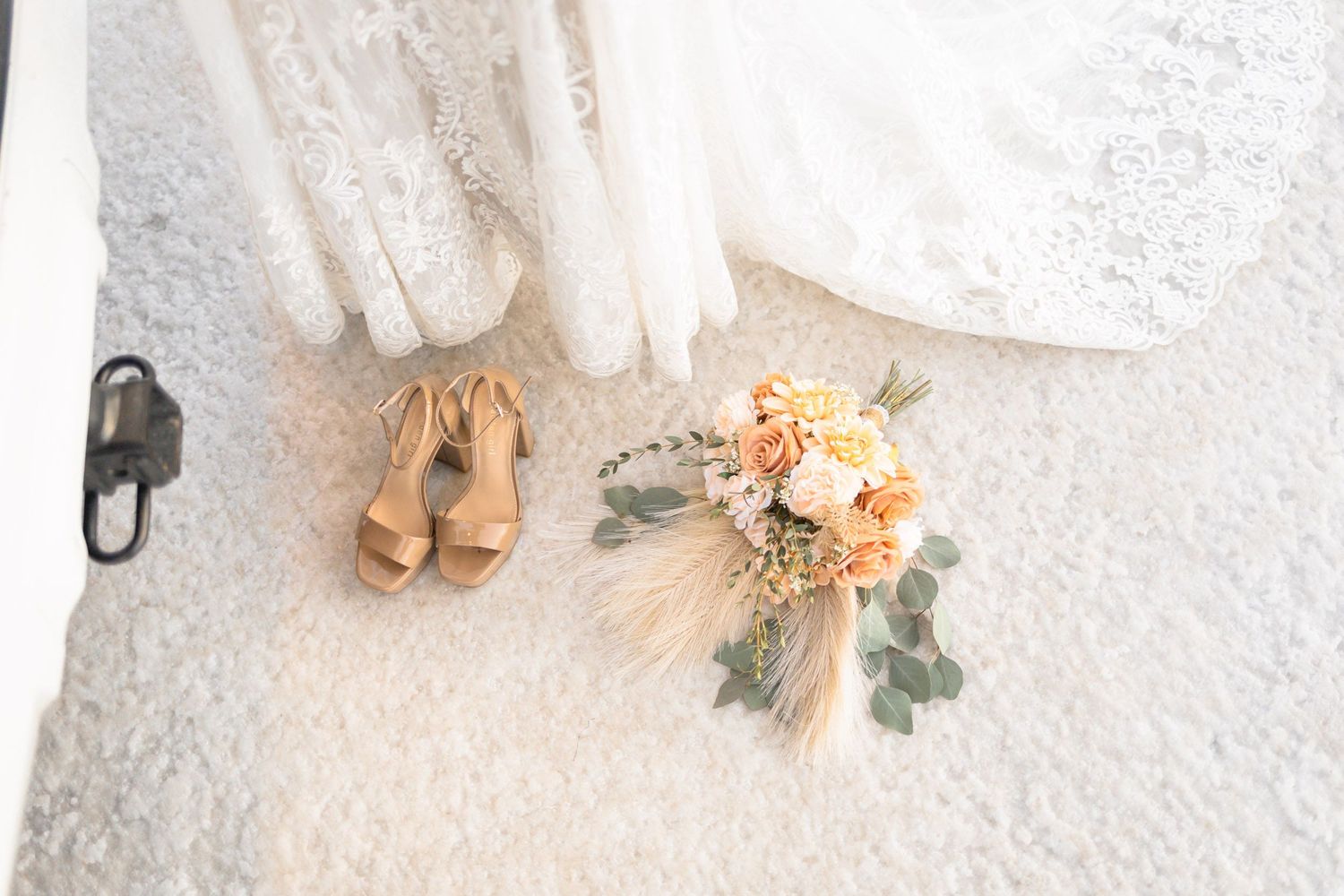 A bridal bouquet with peach roses and eucalyptus lies next to nude wedding shoes and white dress on light carpet.