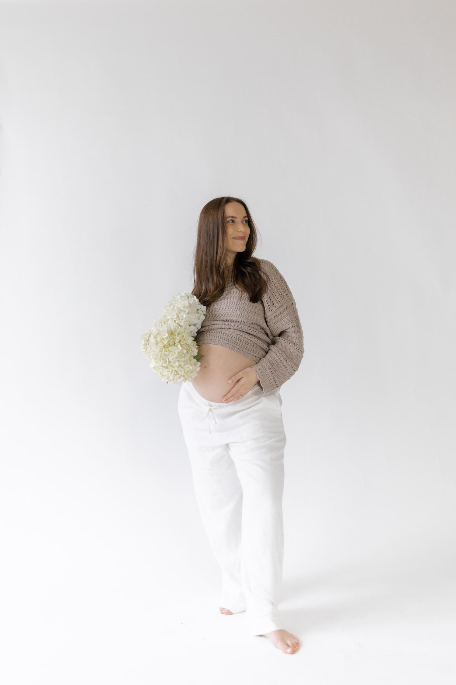 A pregnant woman in a beige crop sweater and white pants holds white hydrangeas against a clean white studio background.
