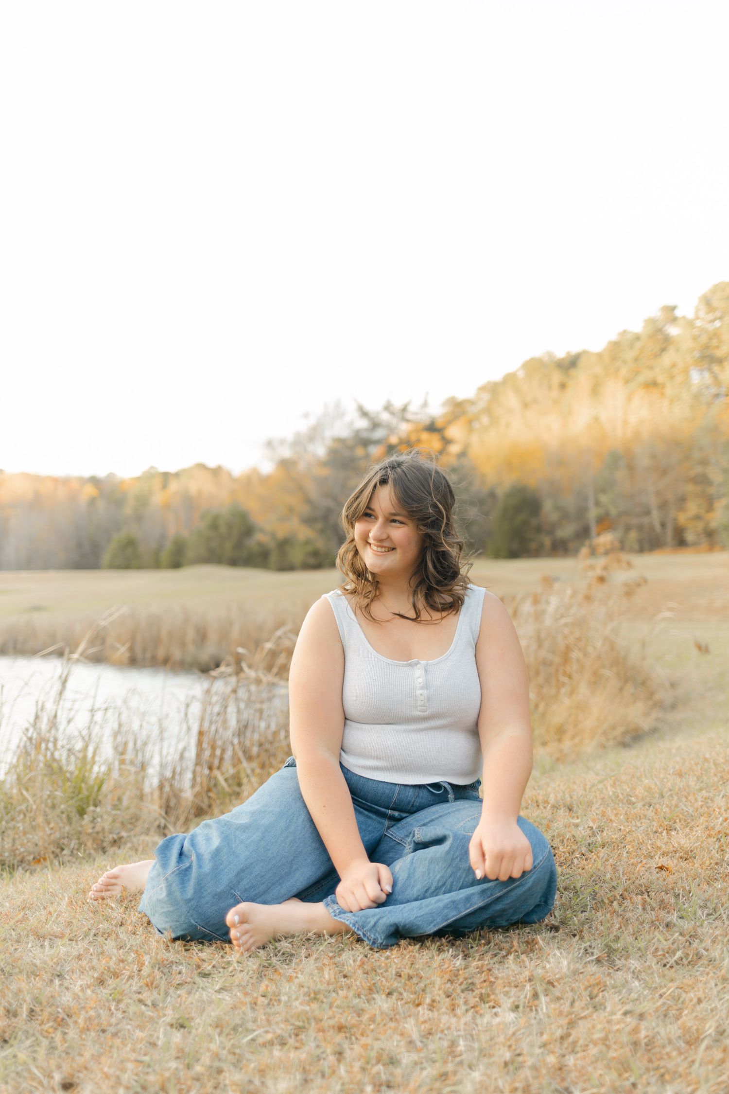 Young woman sitting cross-legged in a golden field near a pond, wearing jeans and a white tank top, smiling outdoors.