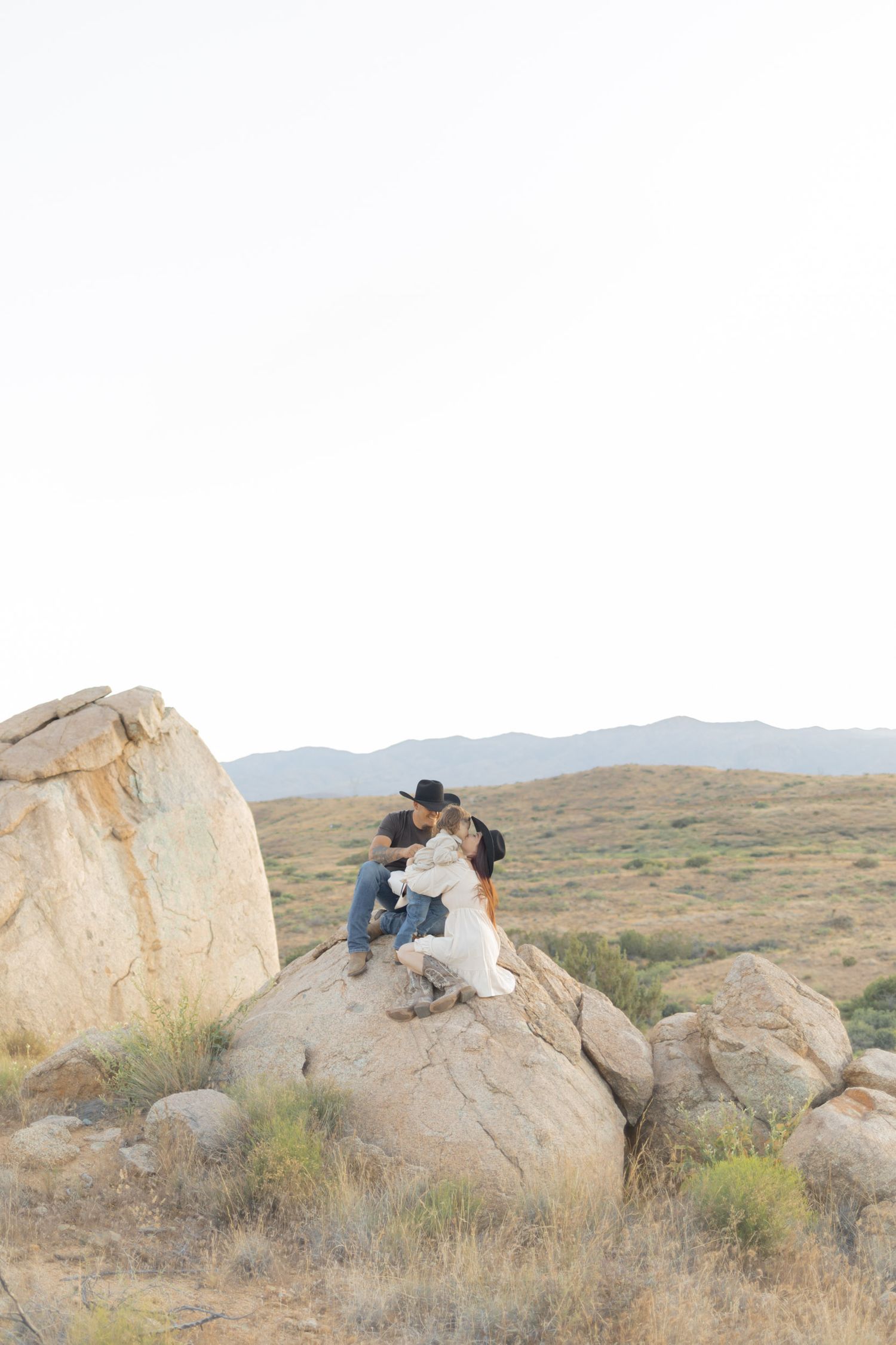 A romantic couple shares a kiss while sitting atop large boulders in a scenic desert landscape.