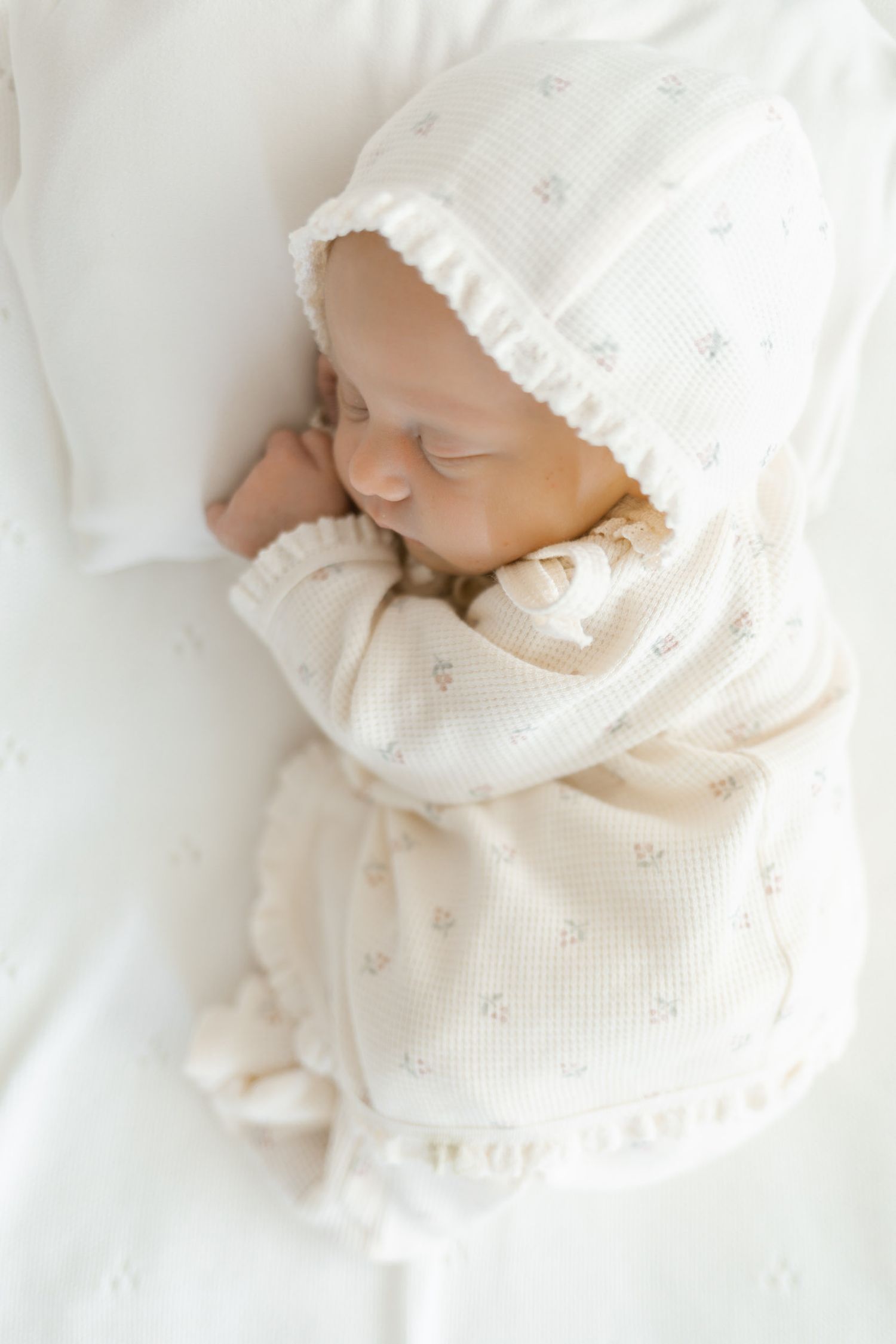 Newborn baby sleeping in cream knit outfit and bonnet on white bedding.