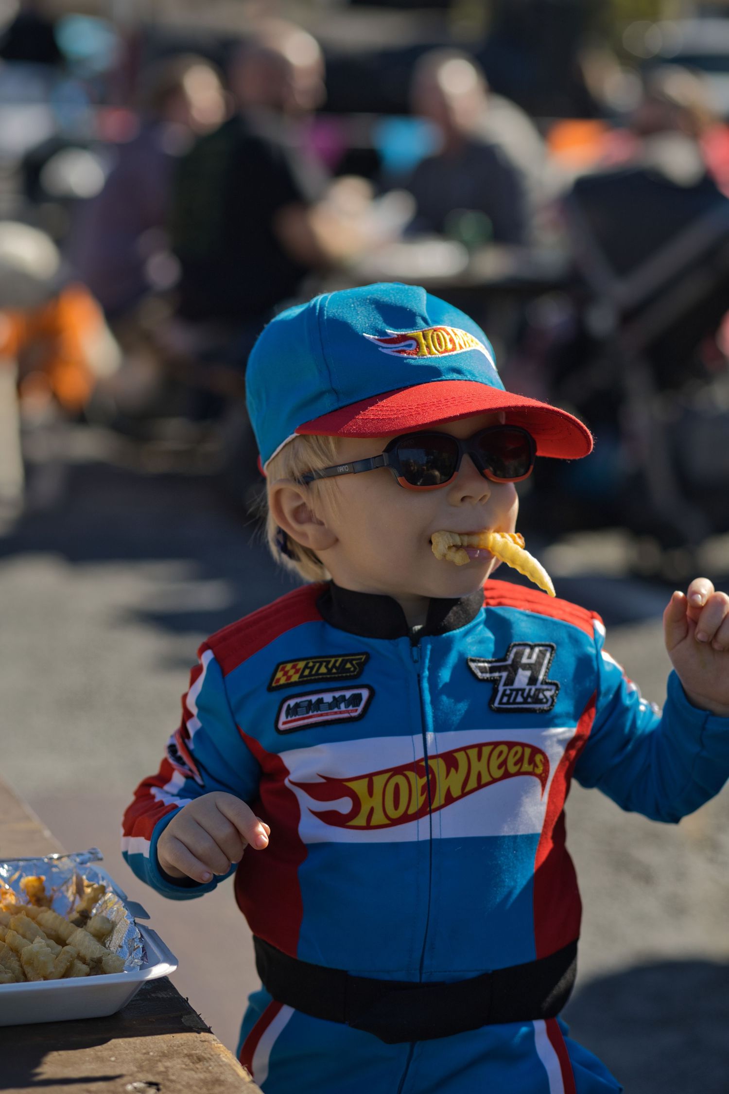 Child in Hot Wheels racing outfit enjoys snacks at outdoor event.