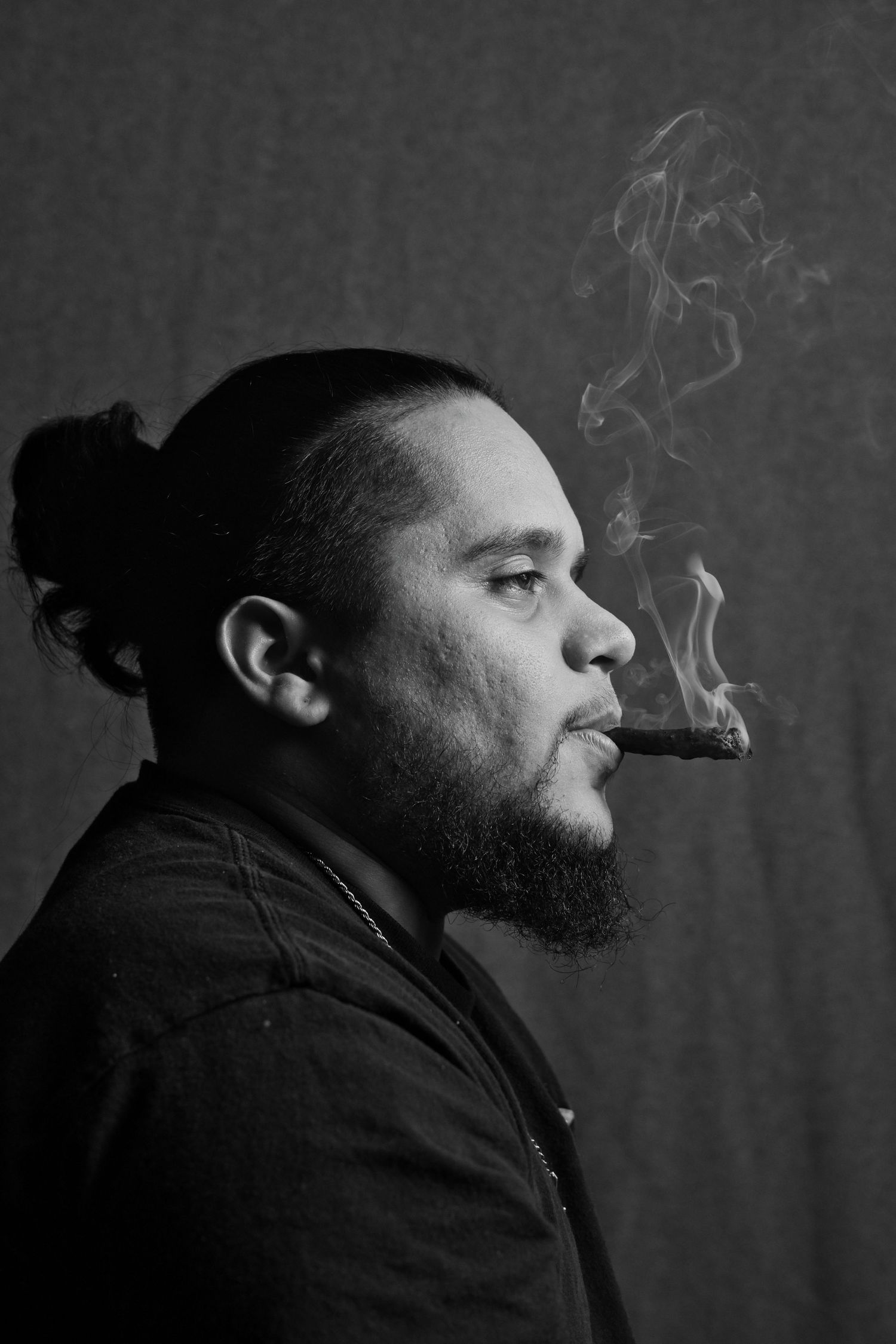 Side profile of bearded man with bun hairstyle smoking a cigar in black and white portrait.