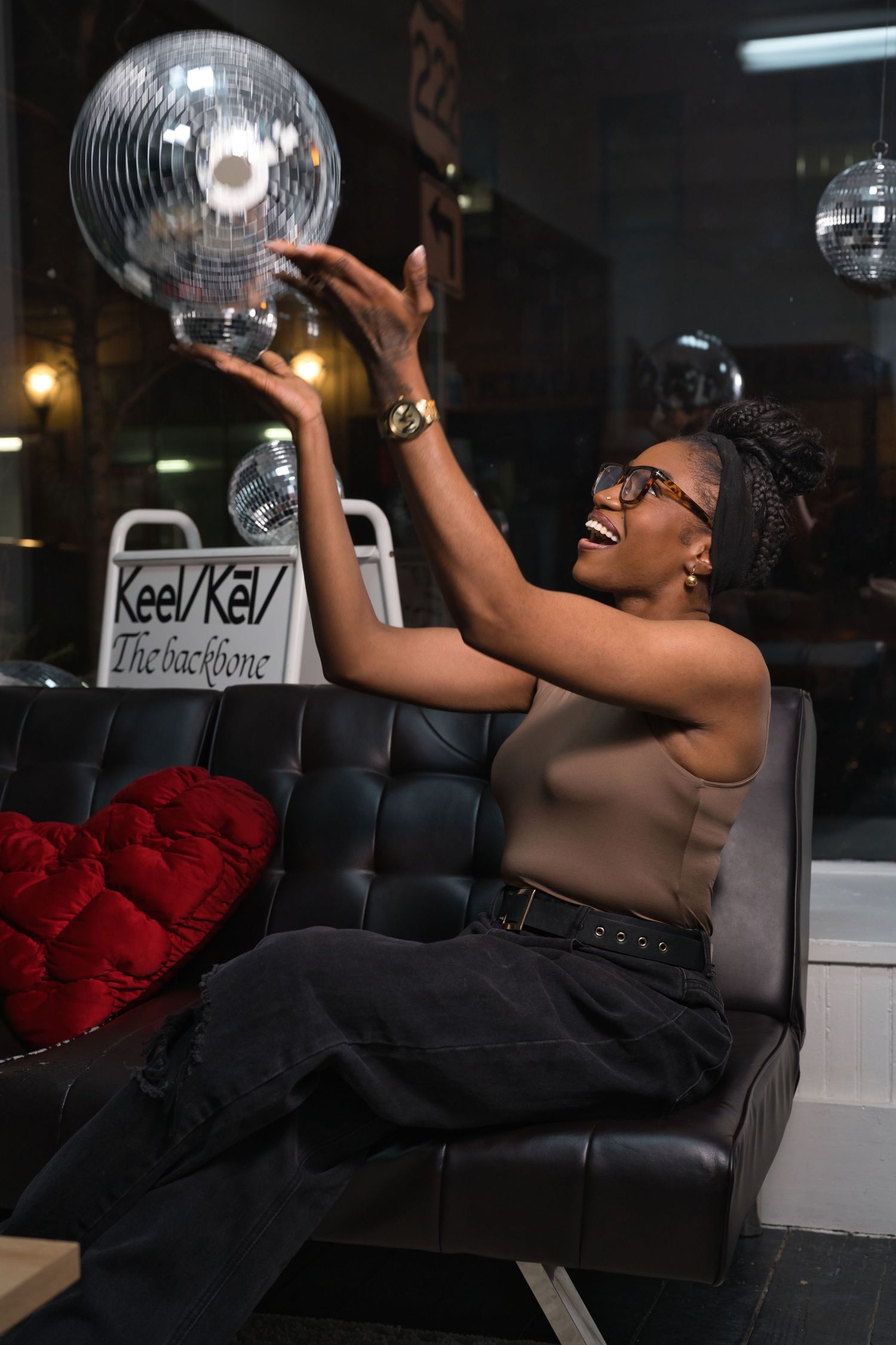A joyful Black woman in glasses holds a disco ball while seated on a leather chair, laughing in a stylish lounge setting.