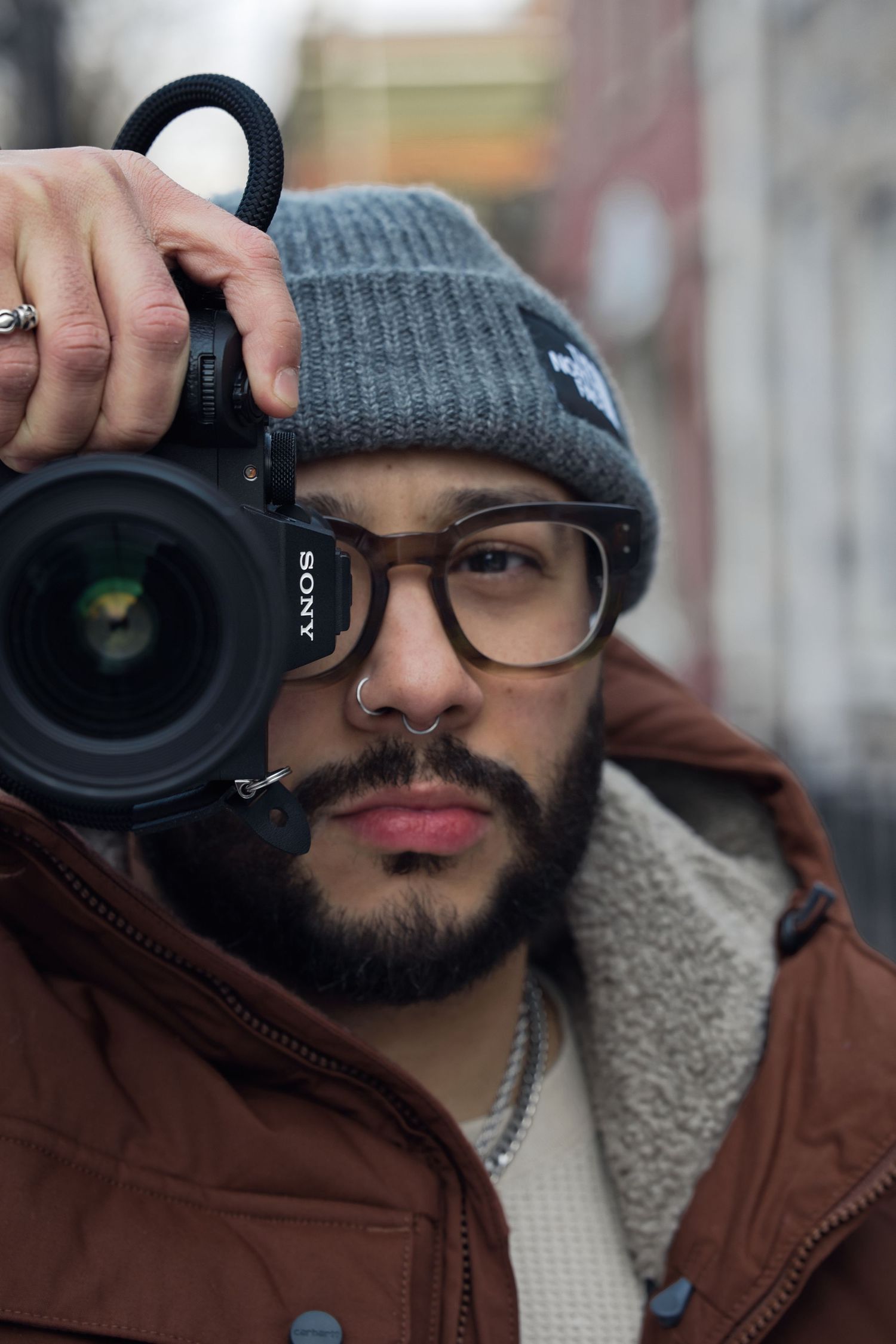 Photographer with beard wearing winter hat and brown jacket holds professional camera.