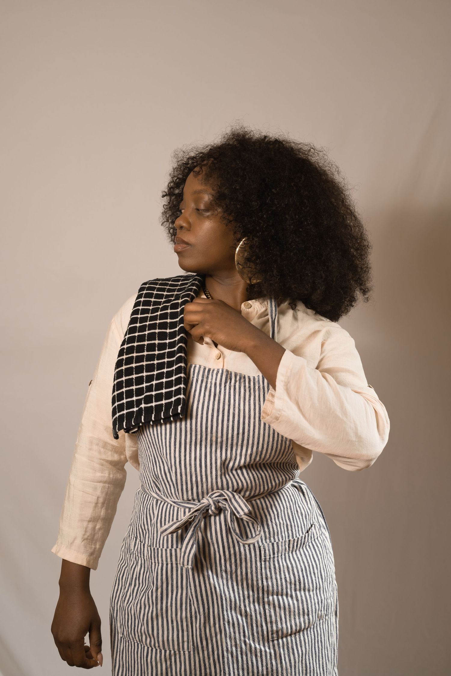 Black woman in striped apron and cream blouse holds checkered cloth, posing against neutral background.