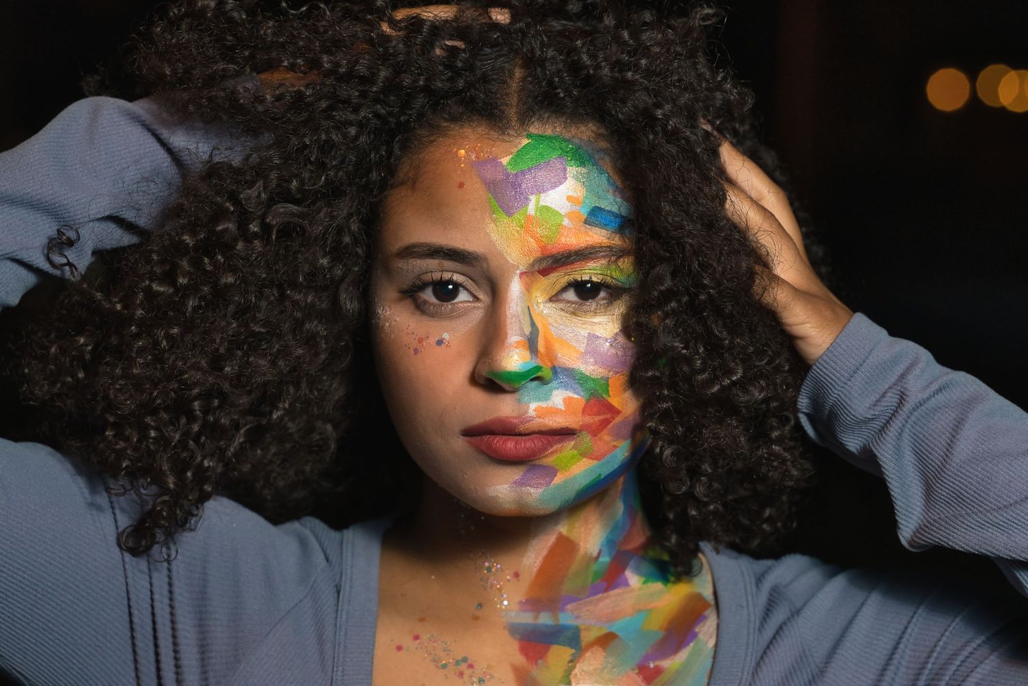Woman with colorful paint art on her face and curly hair poses against a dark background.