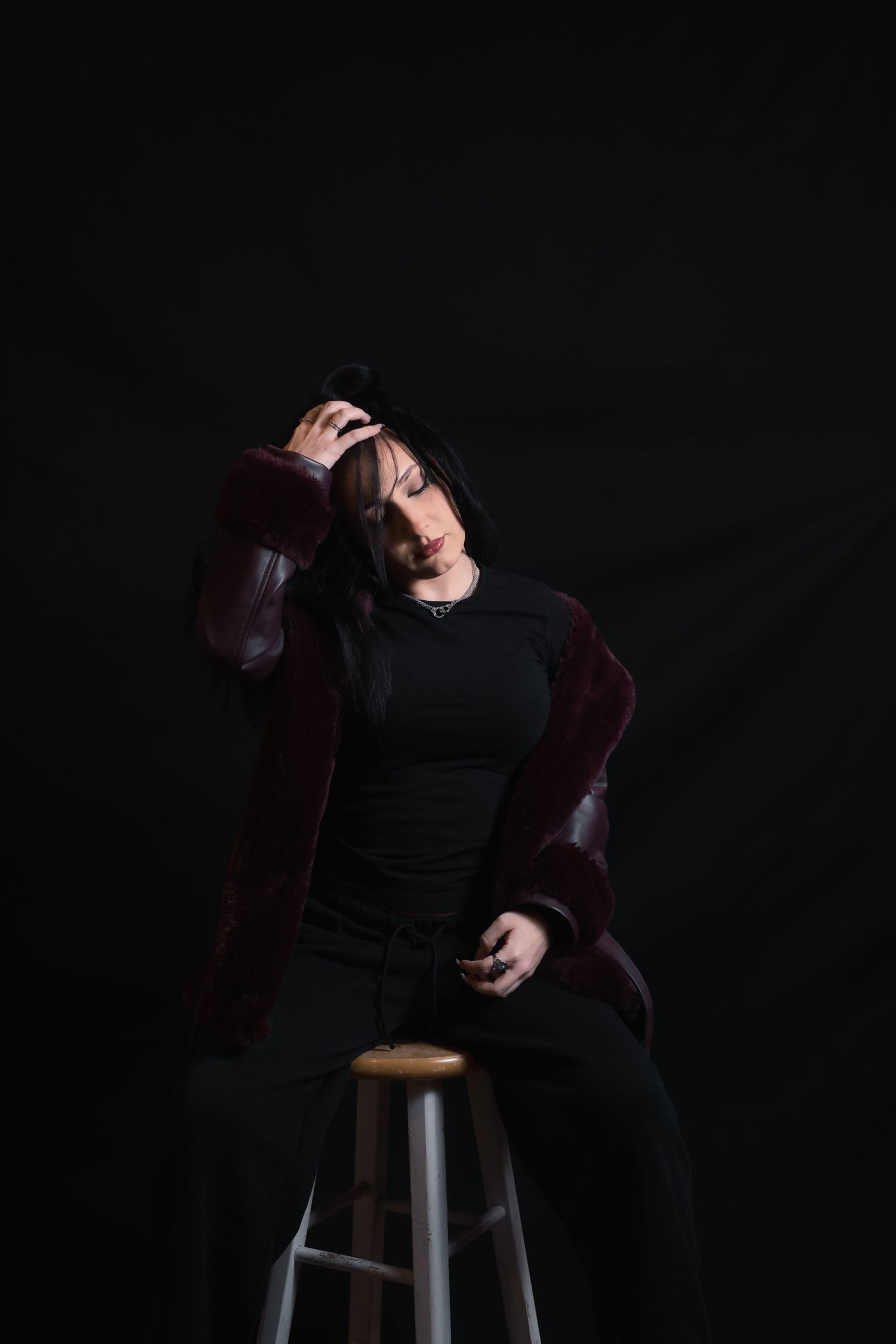 A woman in a black outfit and burgundy jacket sits on a stool, posing dramatically against a dark background.
