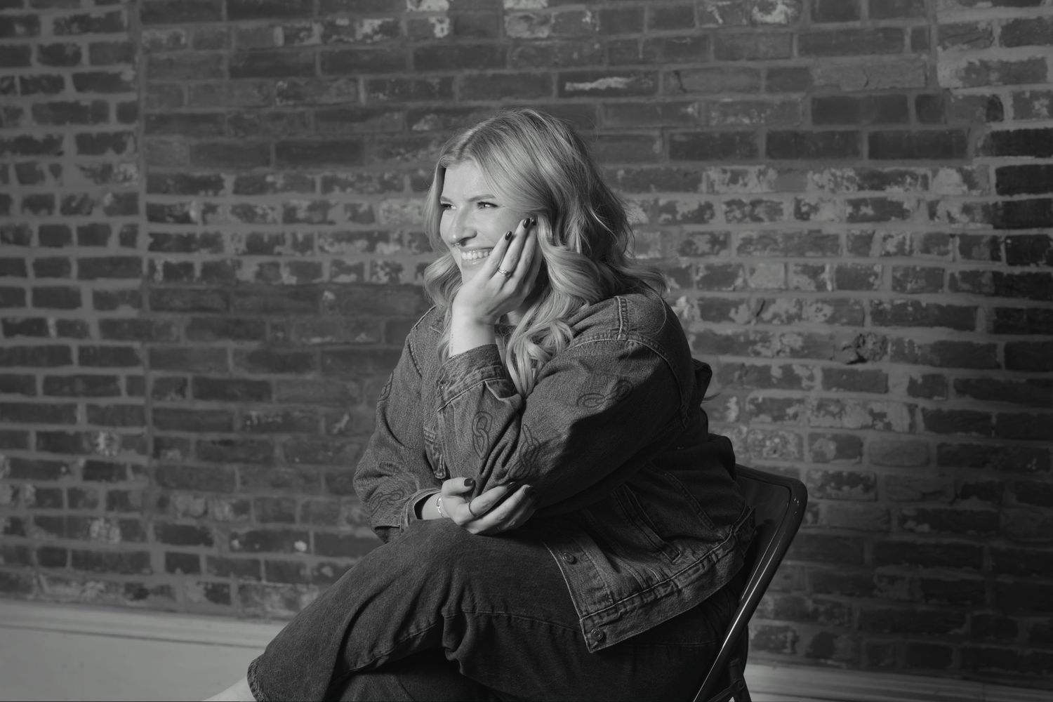 Smiling blonde woman sitting in a chair against a brick wall in a black and white portrait photo.