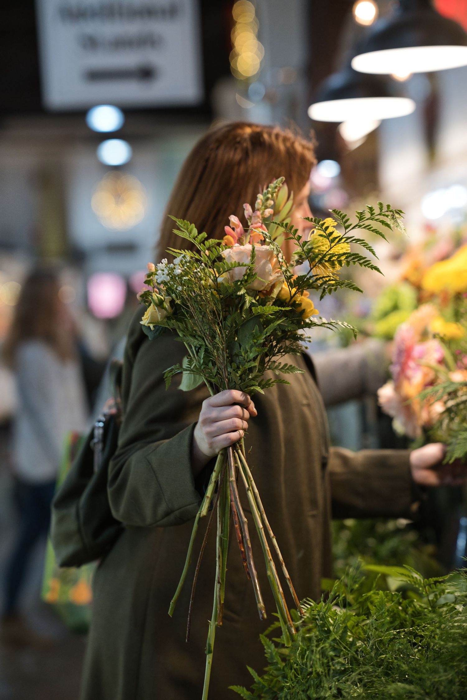 Woman in olive coat holding fresh flower bouquet at a vibrant indoor flower market stall.