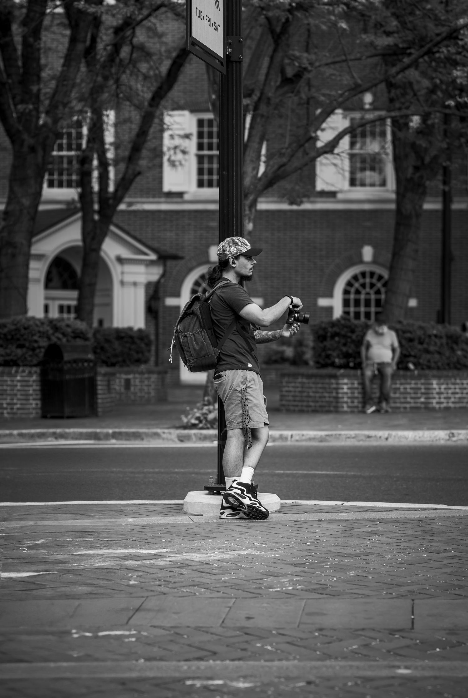 Black and white photo of a man with backpack holding camera on urban street corner with trees and brick building.