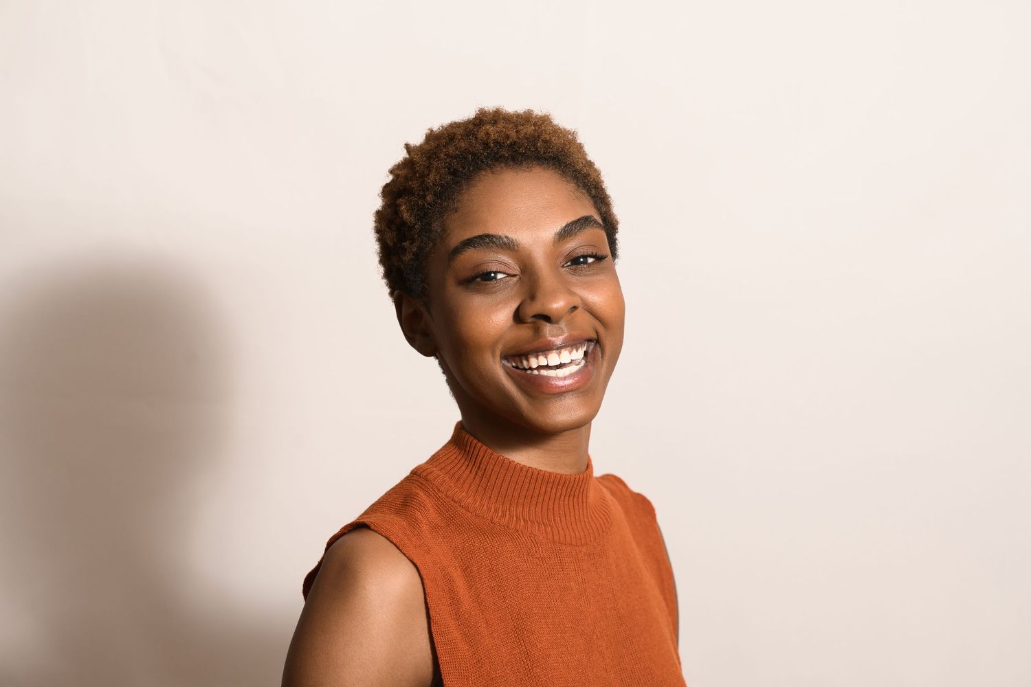 Smiling young Black woman with short natural hair wearing an orange sleeveless top against a light background.