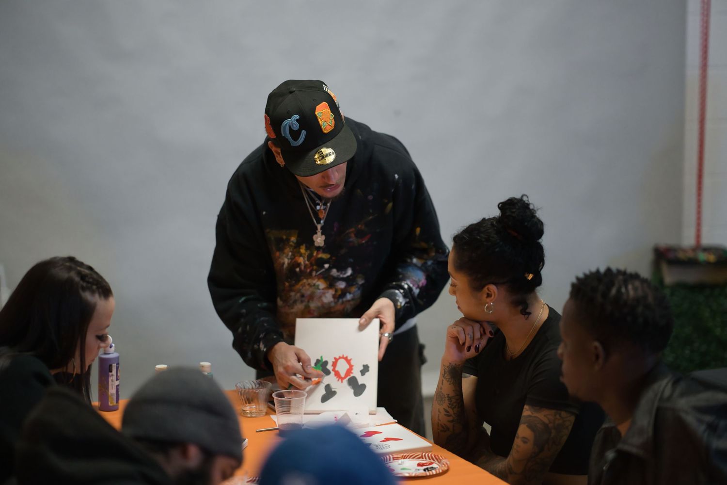 A man in a black hoodie and cap signs merchandise at a table surrounded by fans indoors.