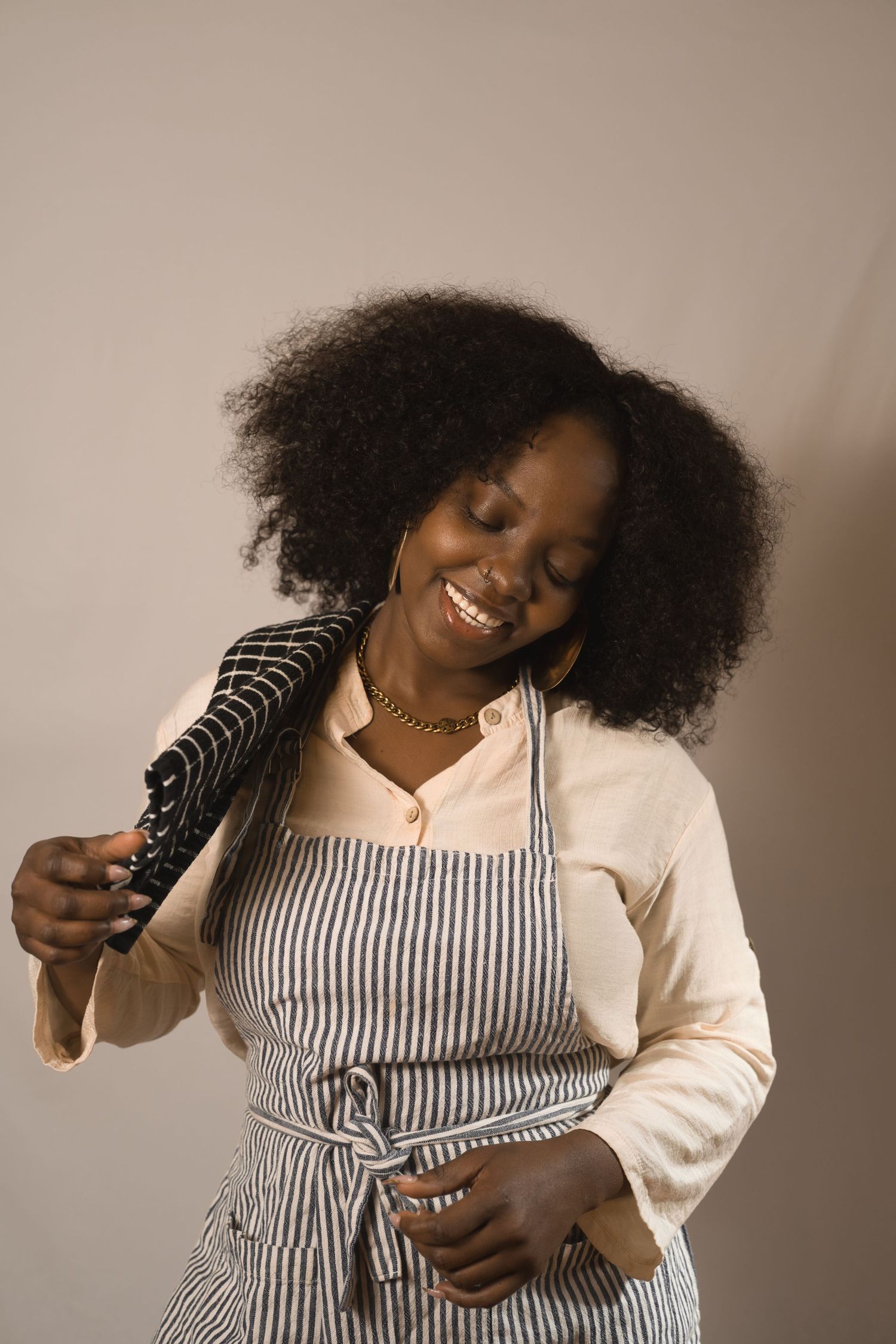 Smiling Black woman with natural hair wearing a striped apron over a cream blouse, holding a kitchen towel.