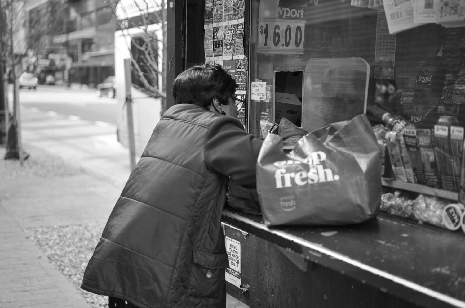 Black and white photo of a person leaning over a street kiosk counter with a fresh grocery bag in an urban setting.