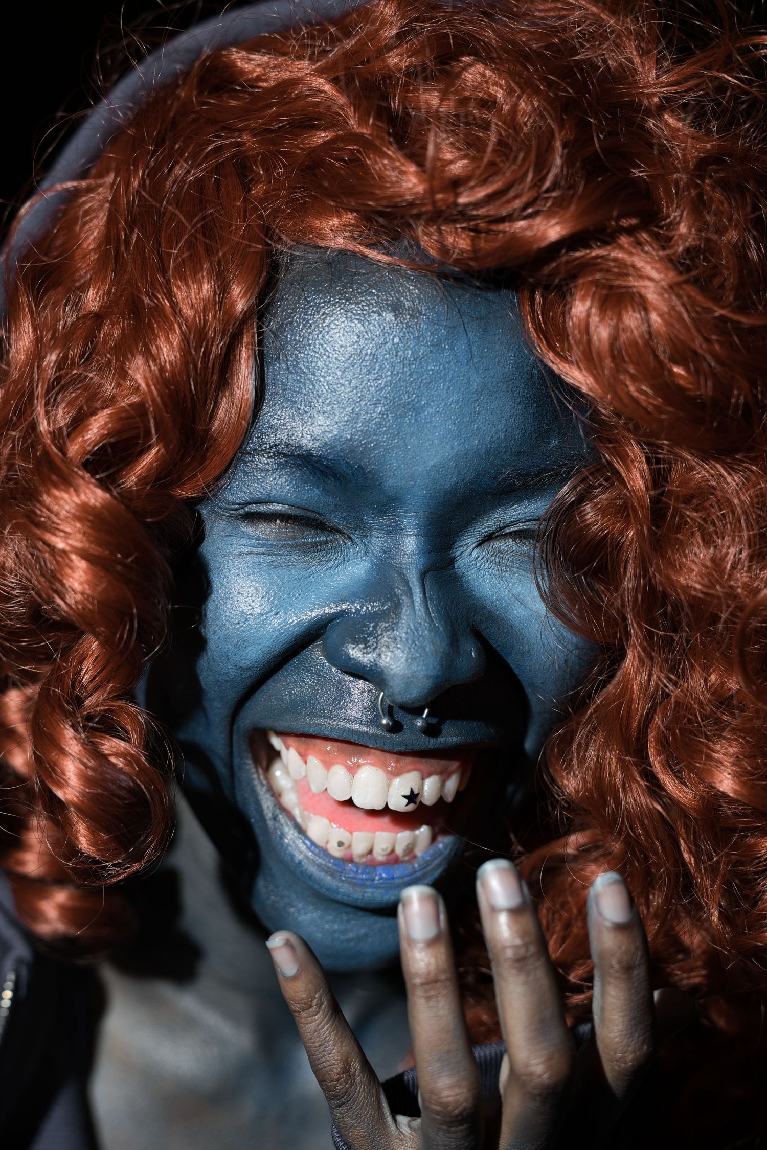 Person with blue face paint and red curly wig laughing joyfully, hands on cheeks, wearing nose ring.