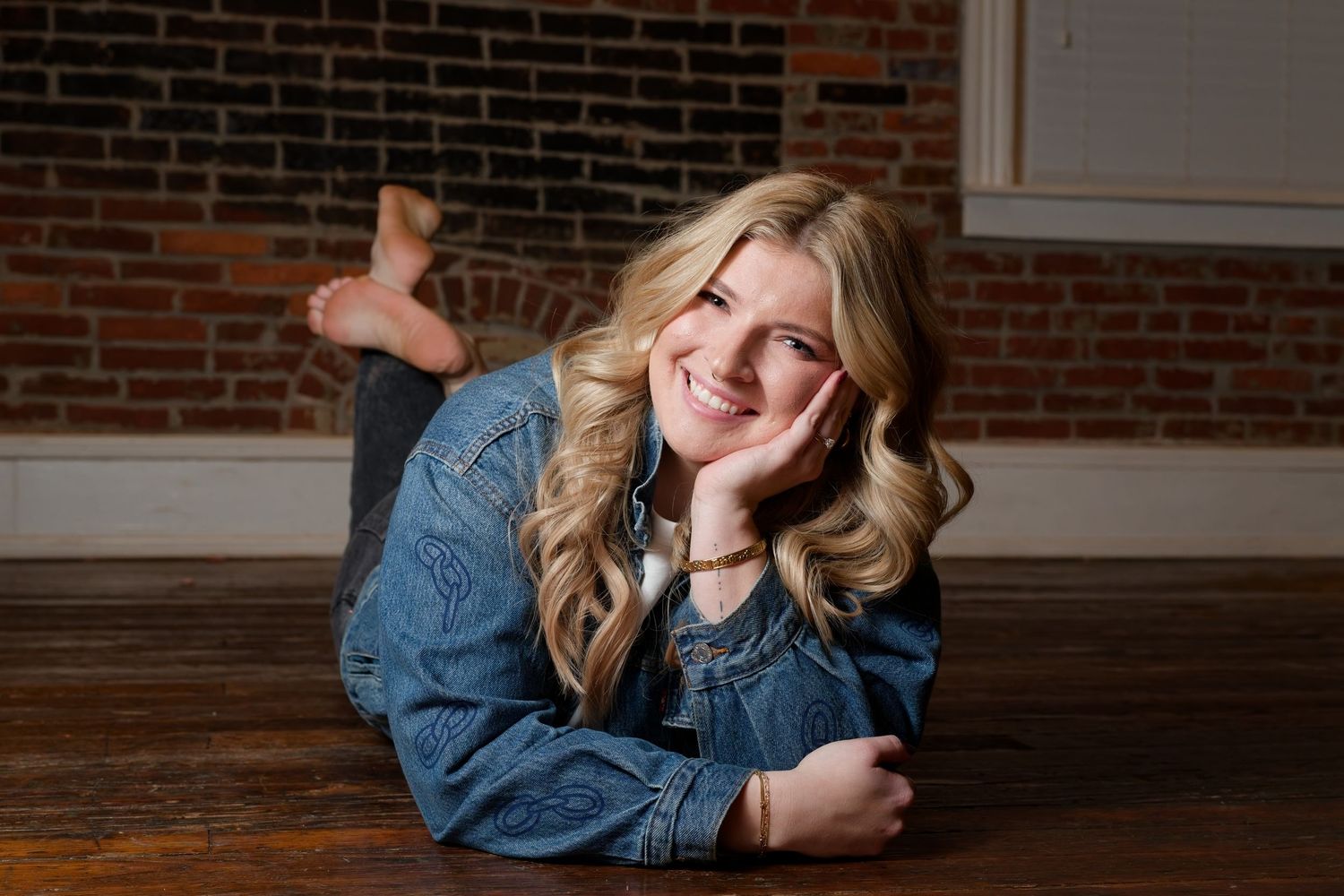 Smiling blonde woman in denim jacket lying on hardwood floor against brick wall.