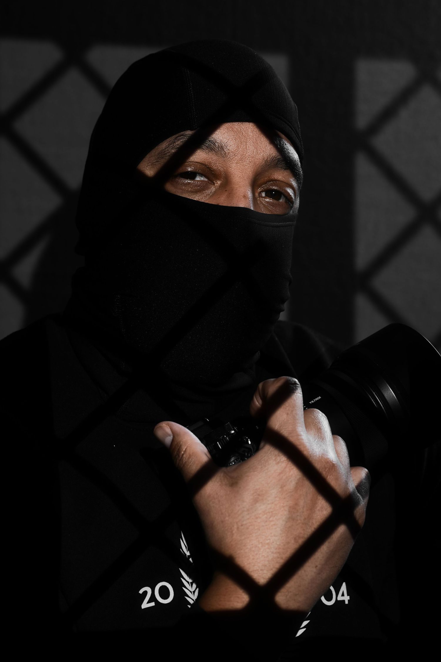 Person in black mask and hoodie holding dumbbells behind a chain-link fence in dramatic lighting.