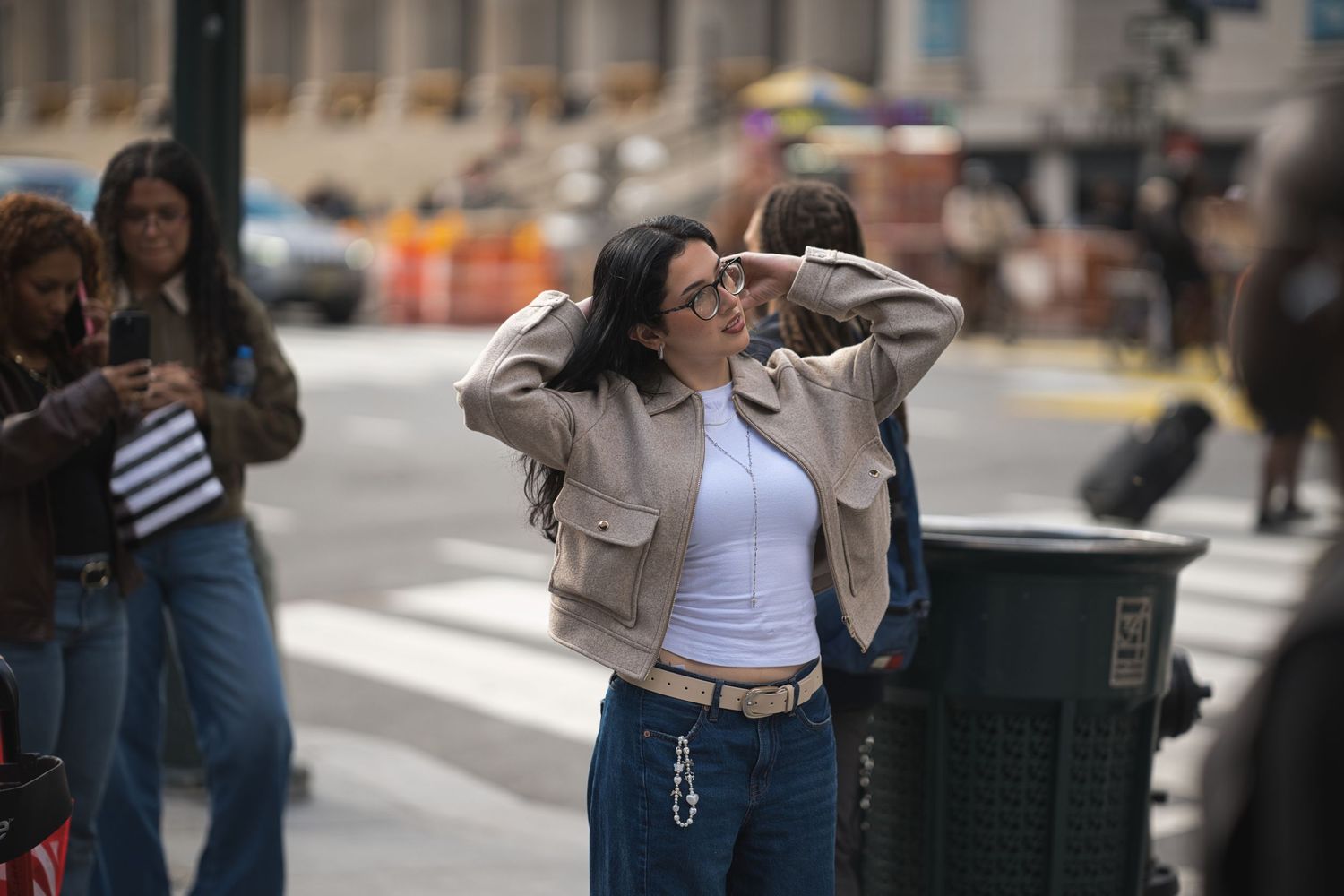 Young woman in beige jacket and jeans posing on busy city street corner.