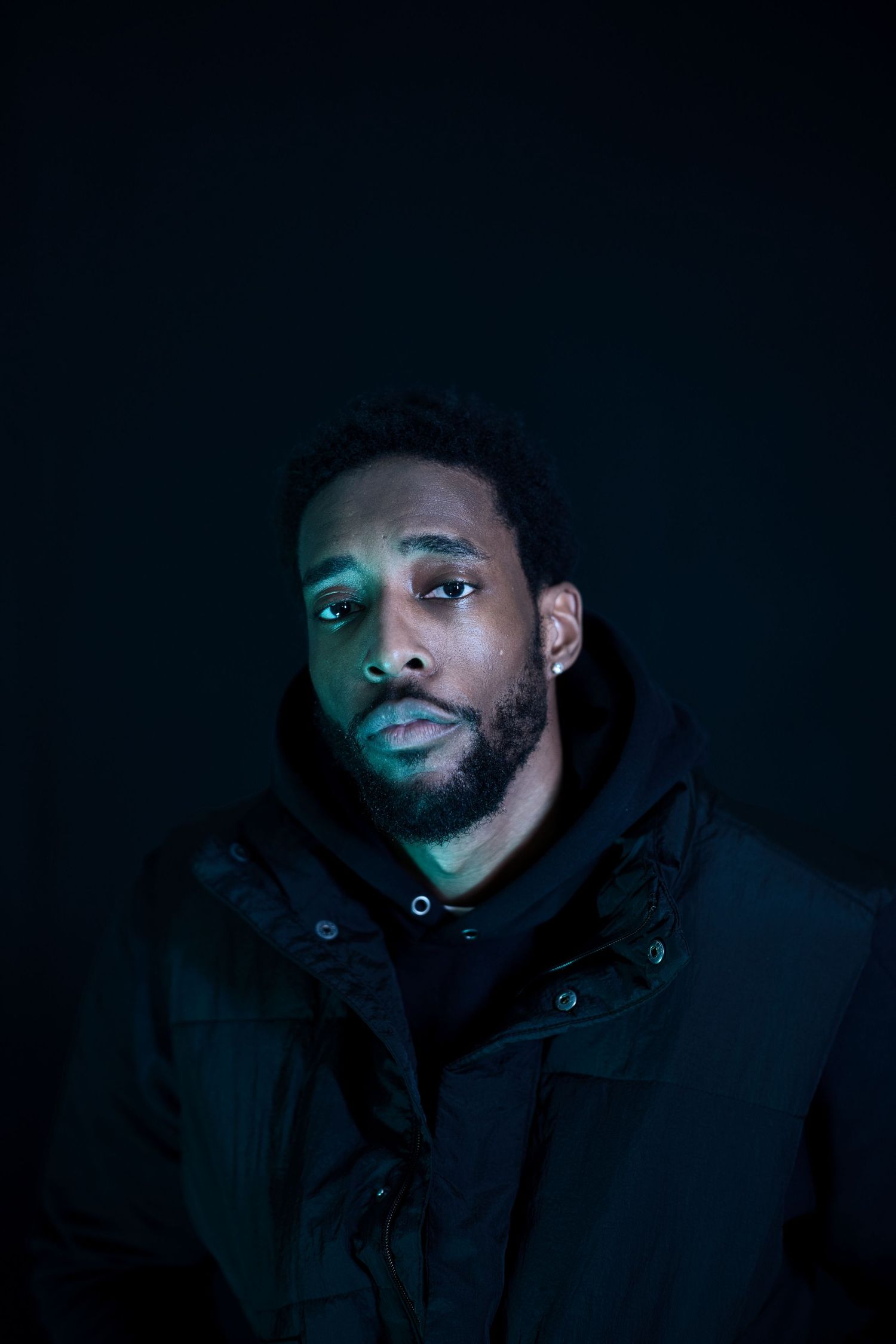 Young Black man with beard wearing black hoodie and jacket, dramatically lit against dark background.