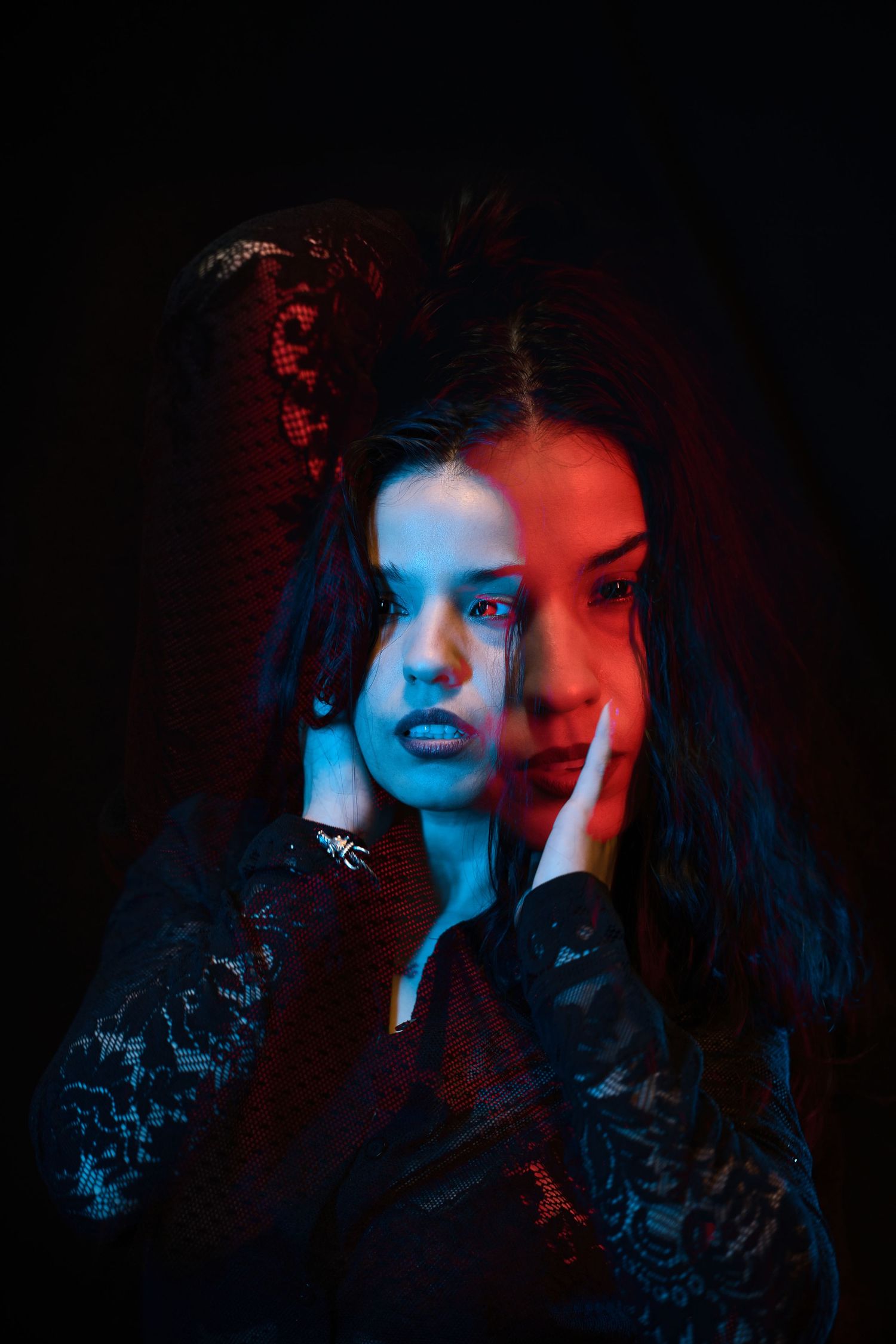 Young woman in black lace dress posed with hands on face under dramatic red and blue neon lighting.