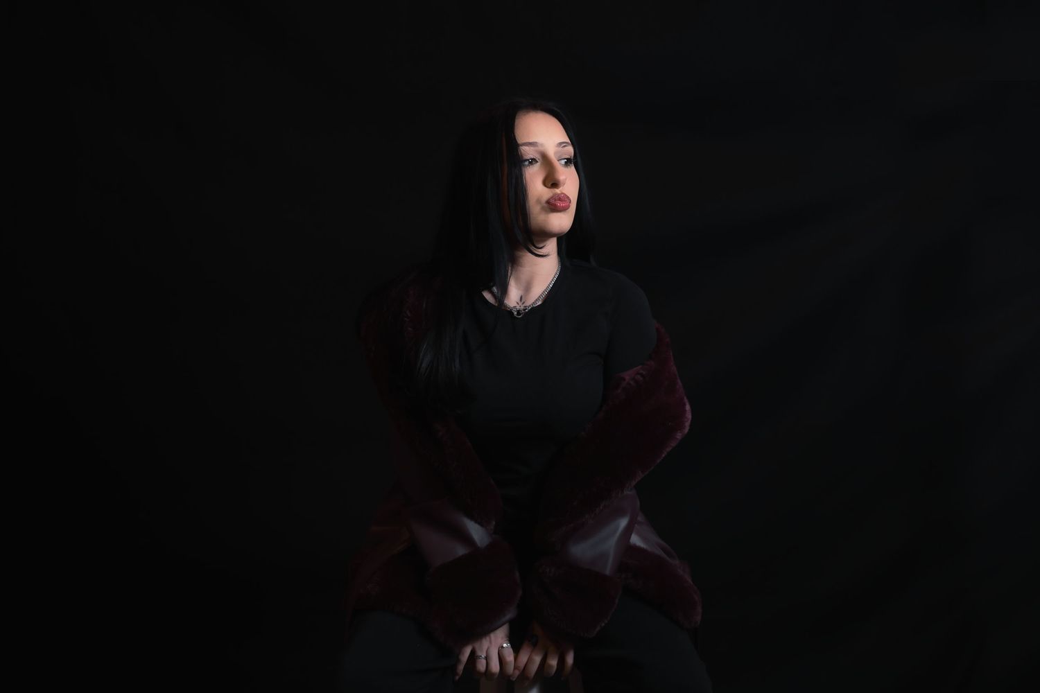 Woman with dark hair in black top and burgundy jacket poses dramatically against dark background.