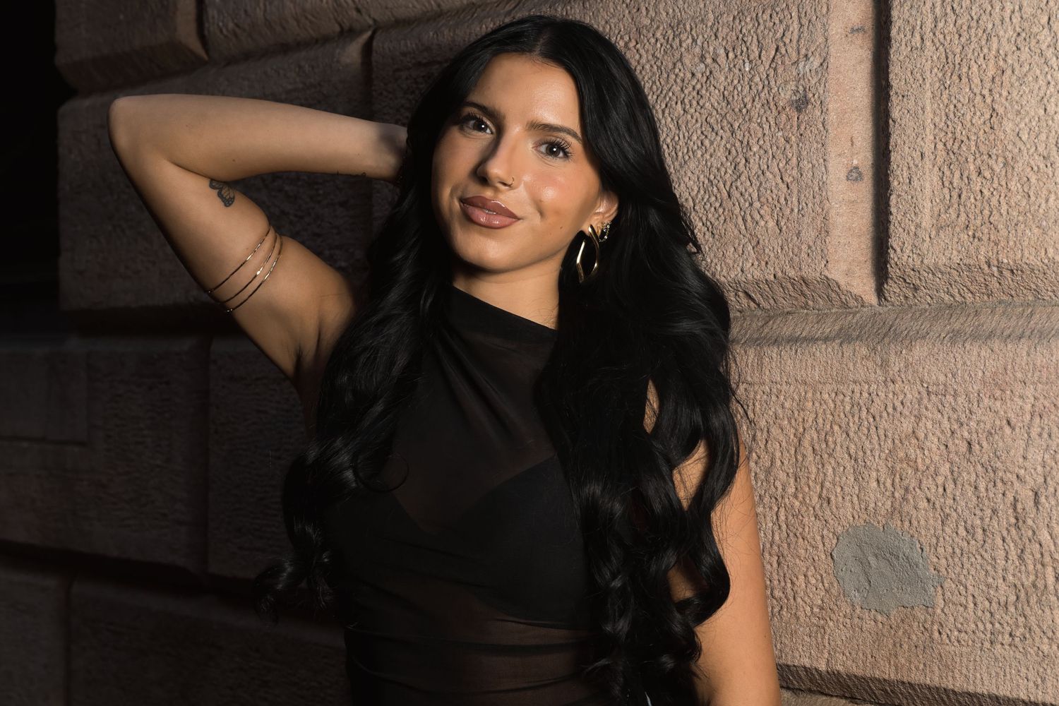 Young woman with long black hair in a black dress poses confidently against a stone wall at night.
