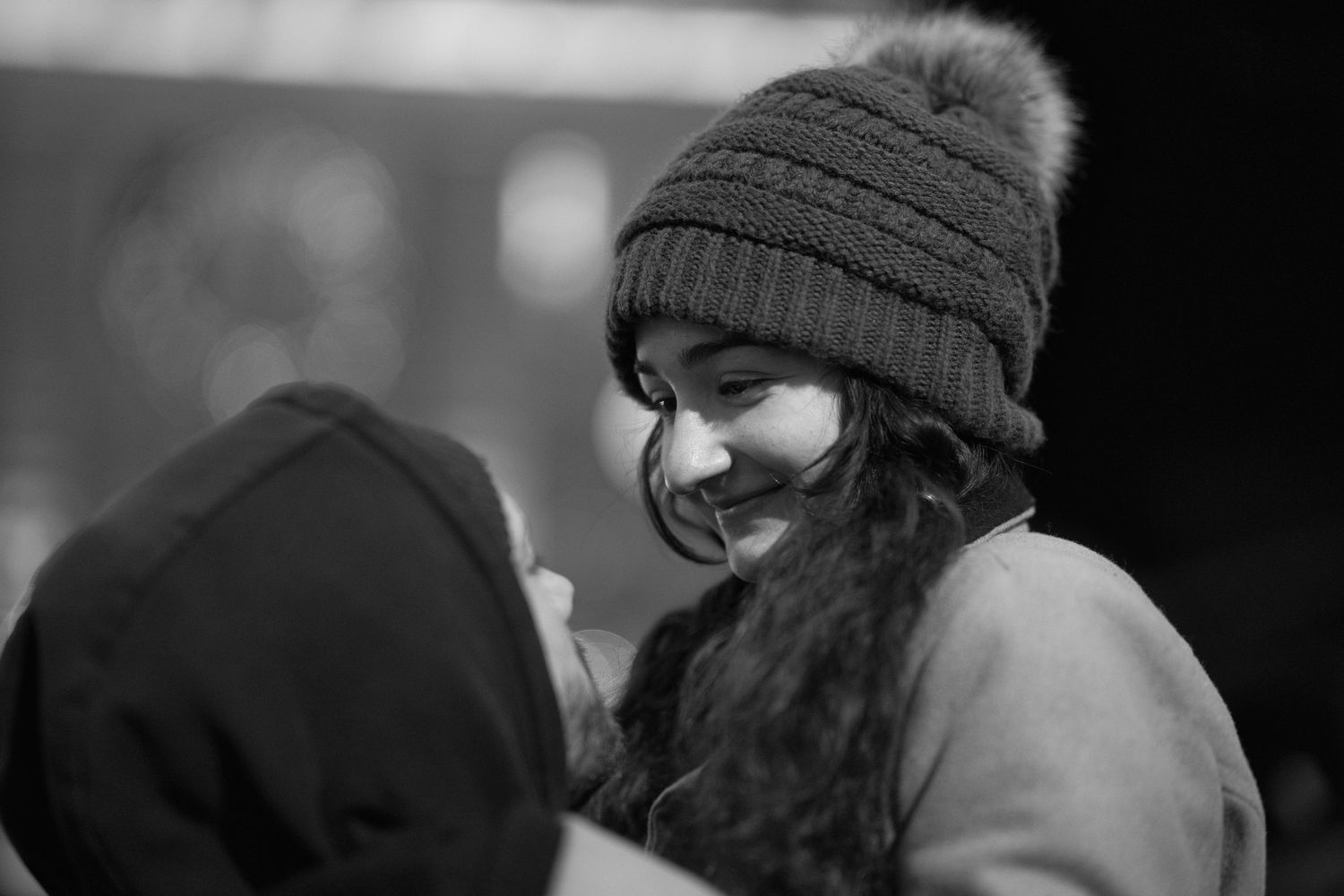 Black and white photo of a smiling young woman wearing a knit pom-pom beanie hat outdoors.