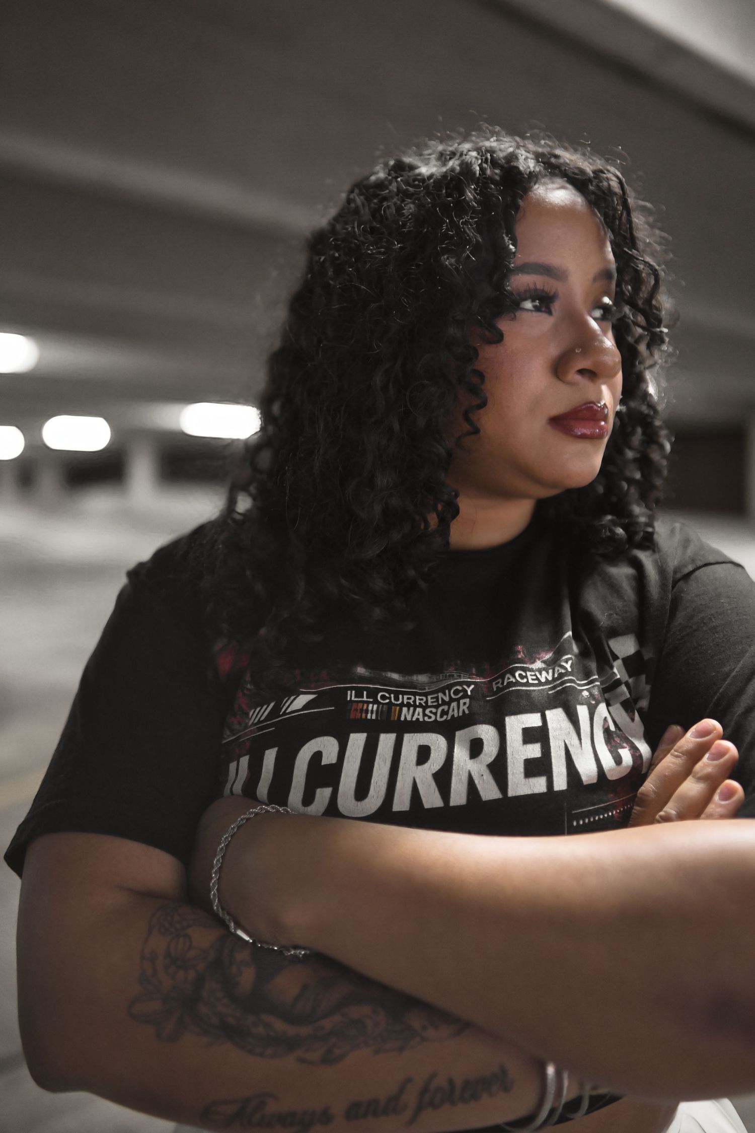 Woman with curly hair wearing Illcurrency t-shirt poses with crossed arms showing tattoos.