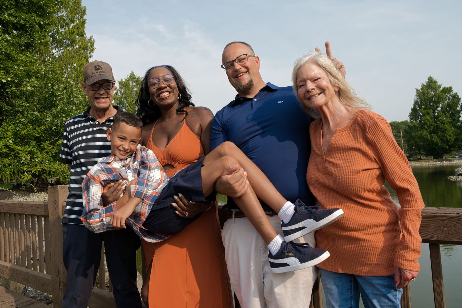 Multigenerational family smiling together outdoors on a bridge, surrounded by lush green trees near a lake.
