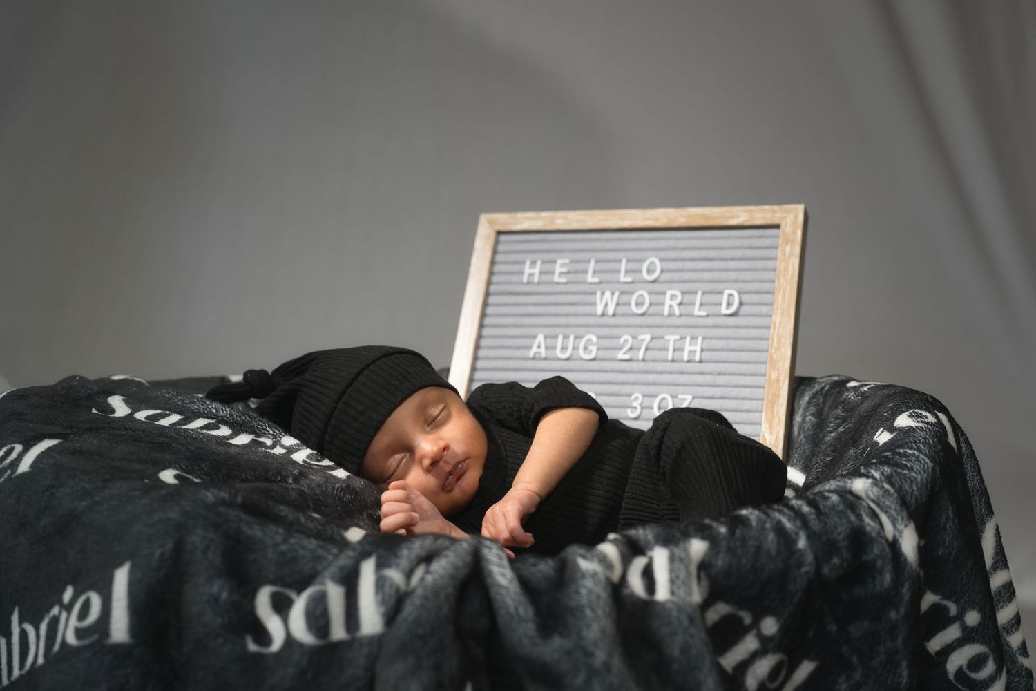 Newborn baby sleeping wrapped in black swaddle beside a Hello World Aug 27th letter board sign.