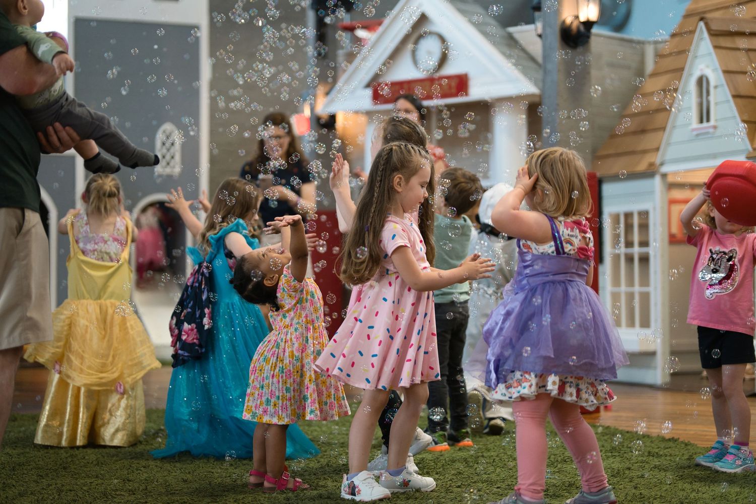 Children playing outdoors in colorful dresses near playhouse structures.