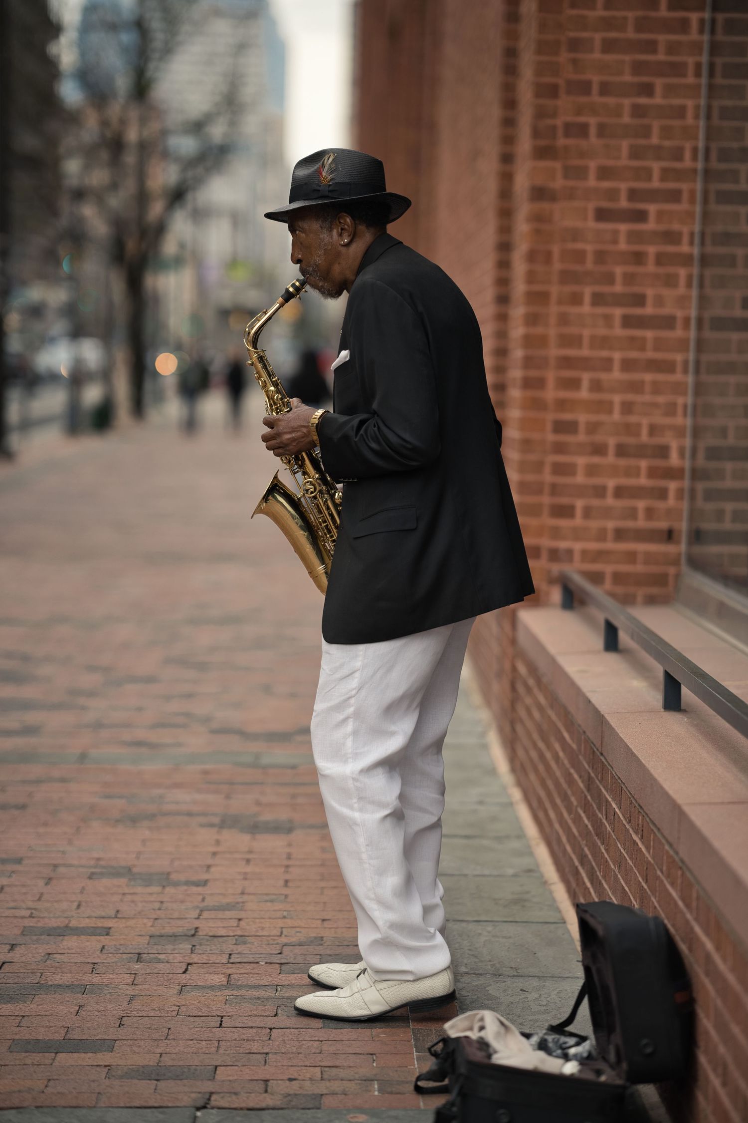 Street musician in black jacket and hat plays saxophone on urban brick sidewalk beside building.