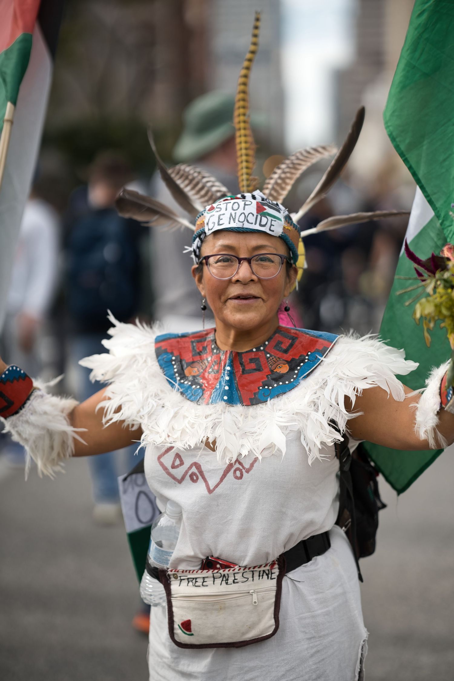 Indigenous woman in traditional regalia with feathered headdress marches in pro-Palestine rally holding flags.