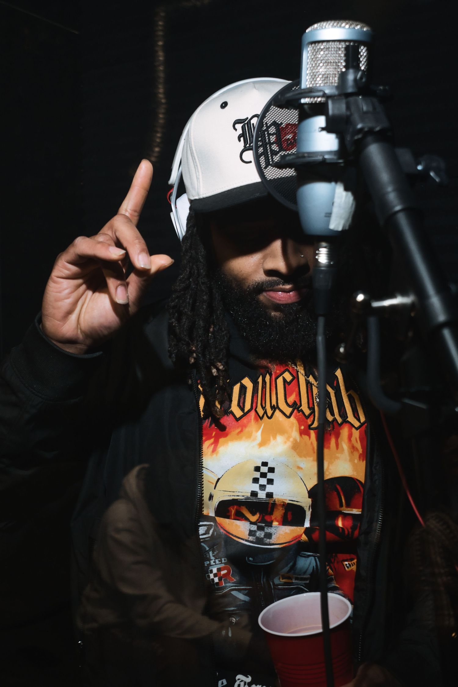 Bearded man with dreadlocks wearing a white helmet cap and graphic tee poses near a studio microphone raising one finger.