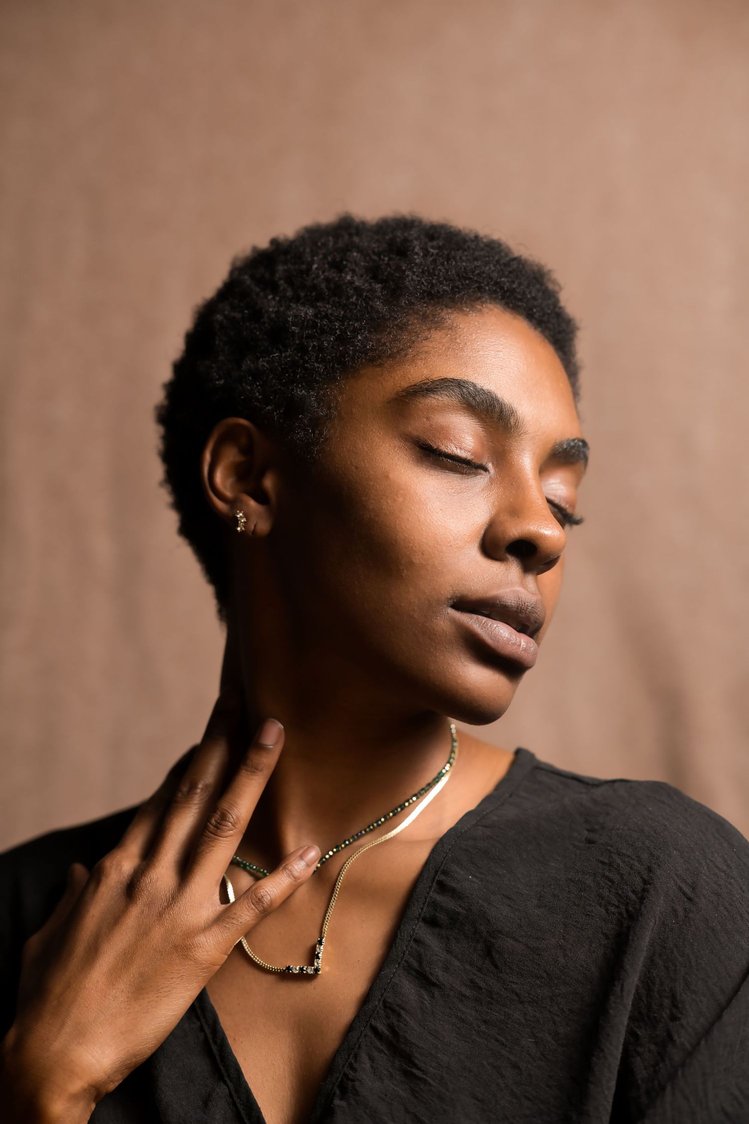 Young Black woman with short natural hair wearing gold jewelry and black top, eyes closed.