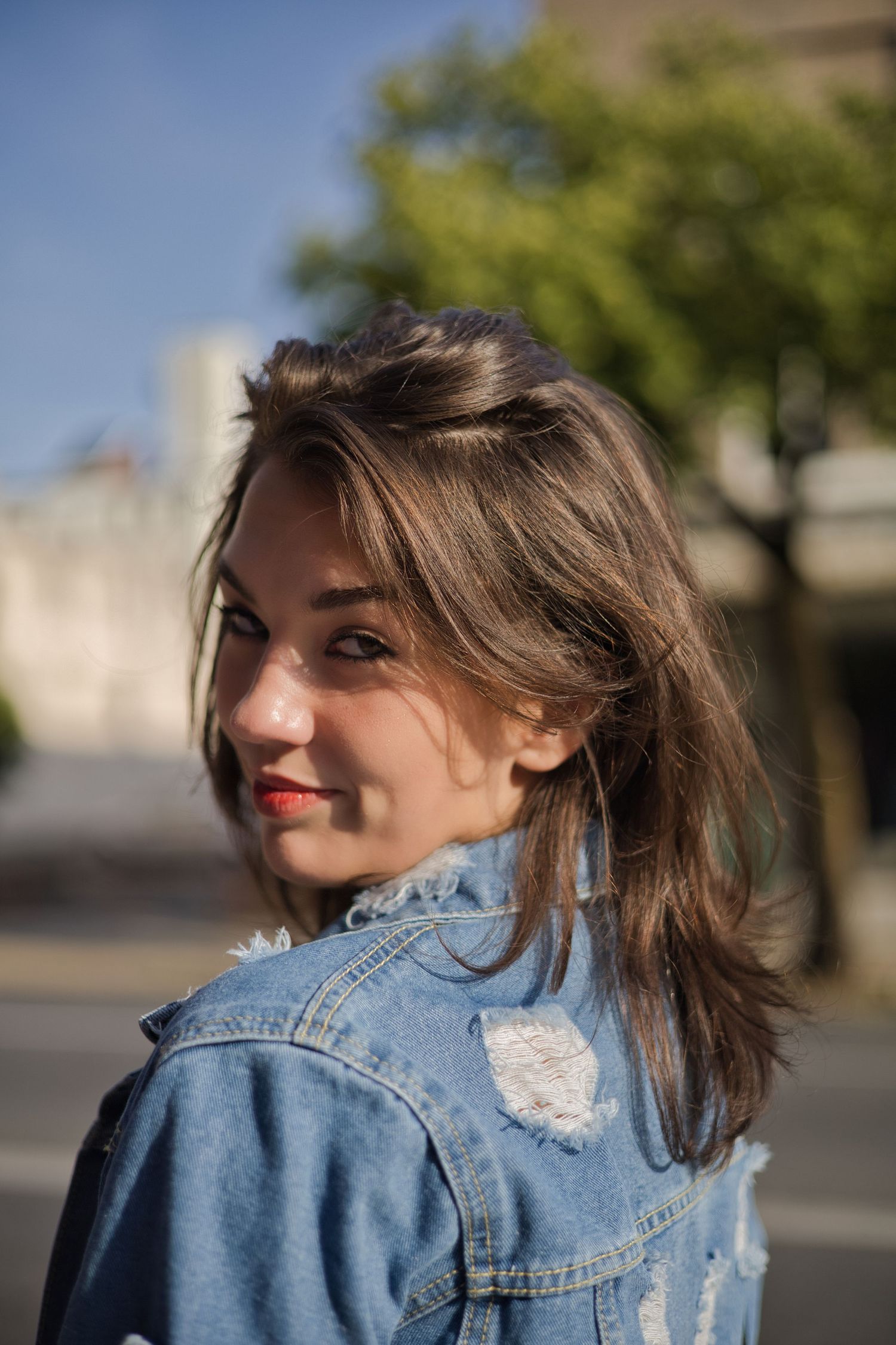 Person in distressed denim jacket looking back over their shoulder outdoors.