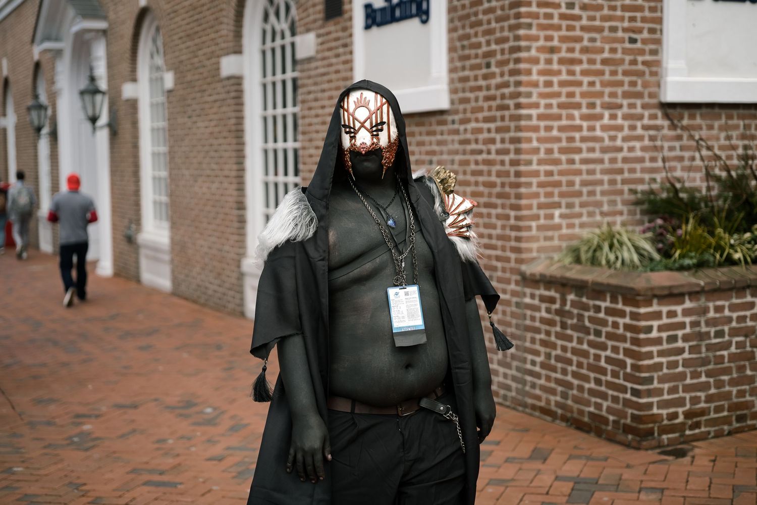 Person in dark villain costume with mask and cape at outdoor convention event.