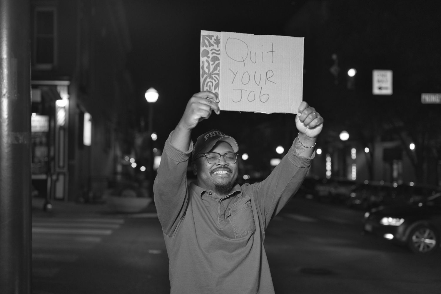 Man holding a cardboard sign reading Quit Your Job on a city street at night in black and white photo.