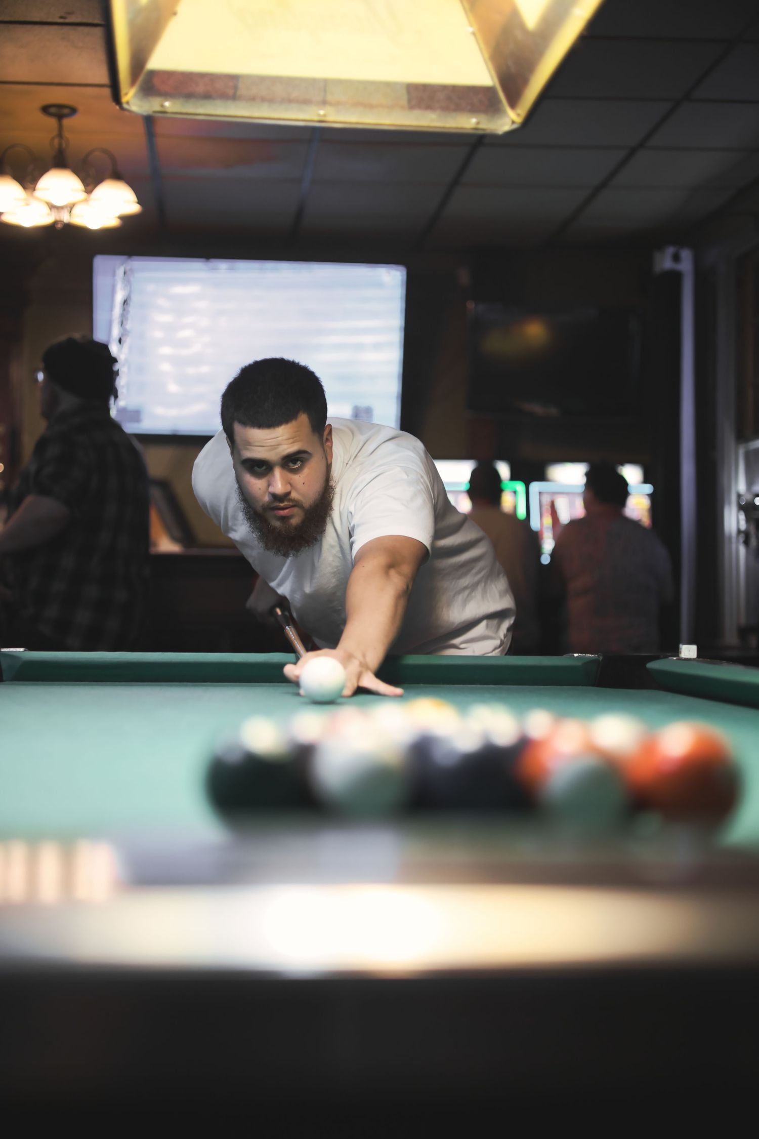 A person in a white t-shirt lines up a shot while playing pool in a dimly lit bar setting.