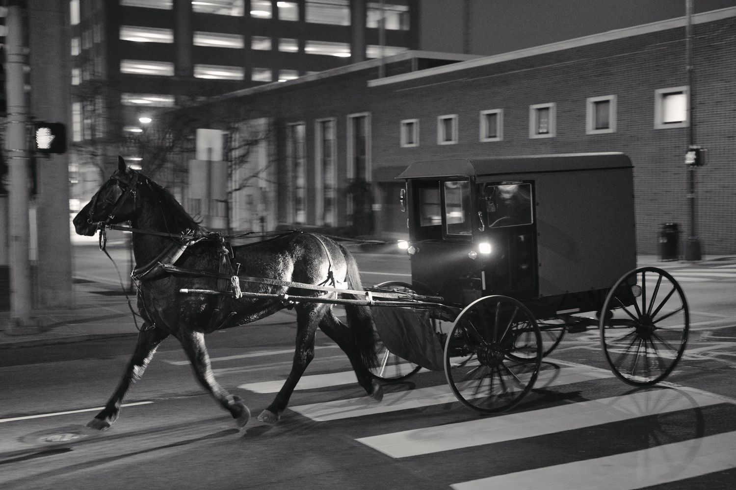 Black and white photo of a horse-drawn Amish buggy trotting through a city street at night.
