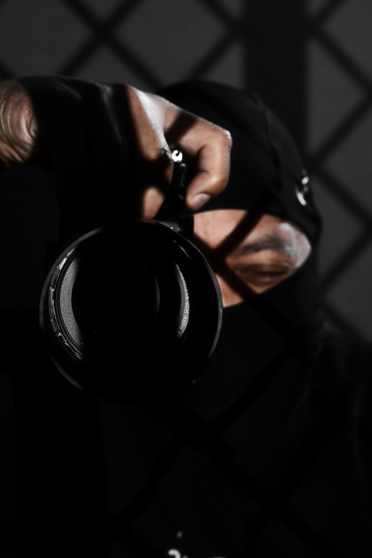 Person in black balaclava holding a professional camera lens pointed forward in dramatic low-key lighting.