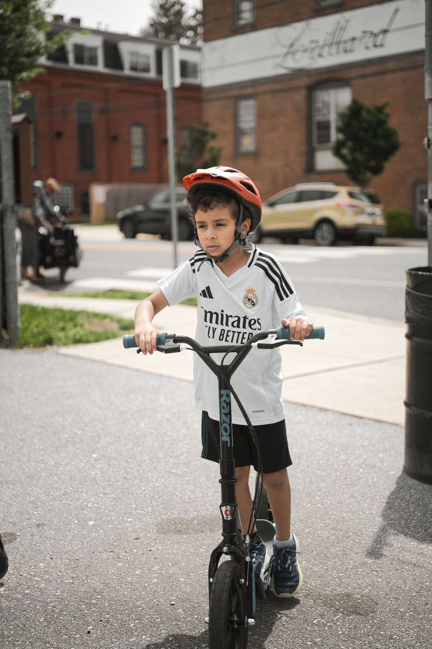 Children riding scooters on residential street wearing safety gear.