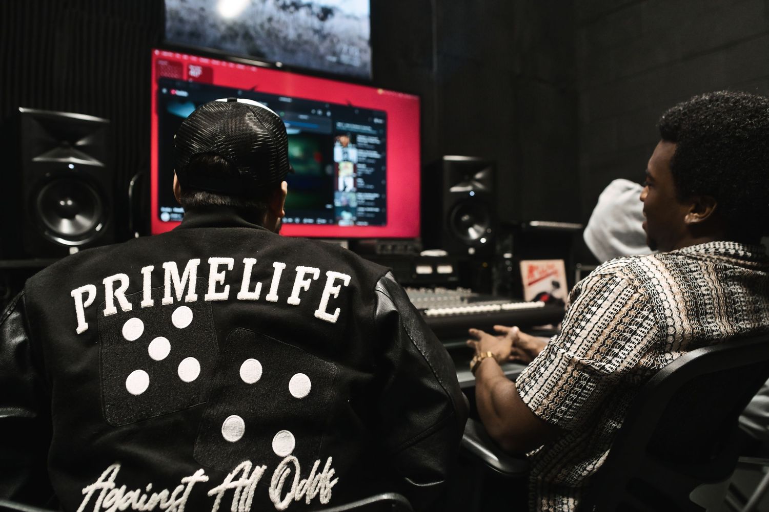 Two men in recording studio with PRIMELIFE jacket visible from behind.