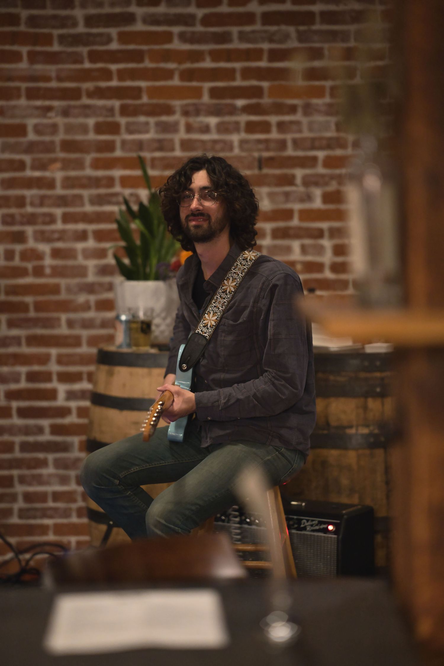 Curly-haired guitarist sitting on a stool in a rustic brick-walled venue, holding an electric guitar with a decorative strap.