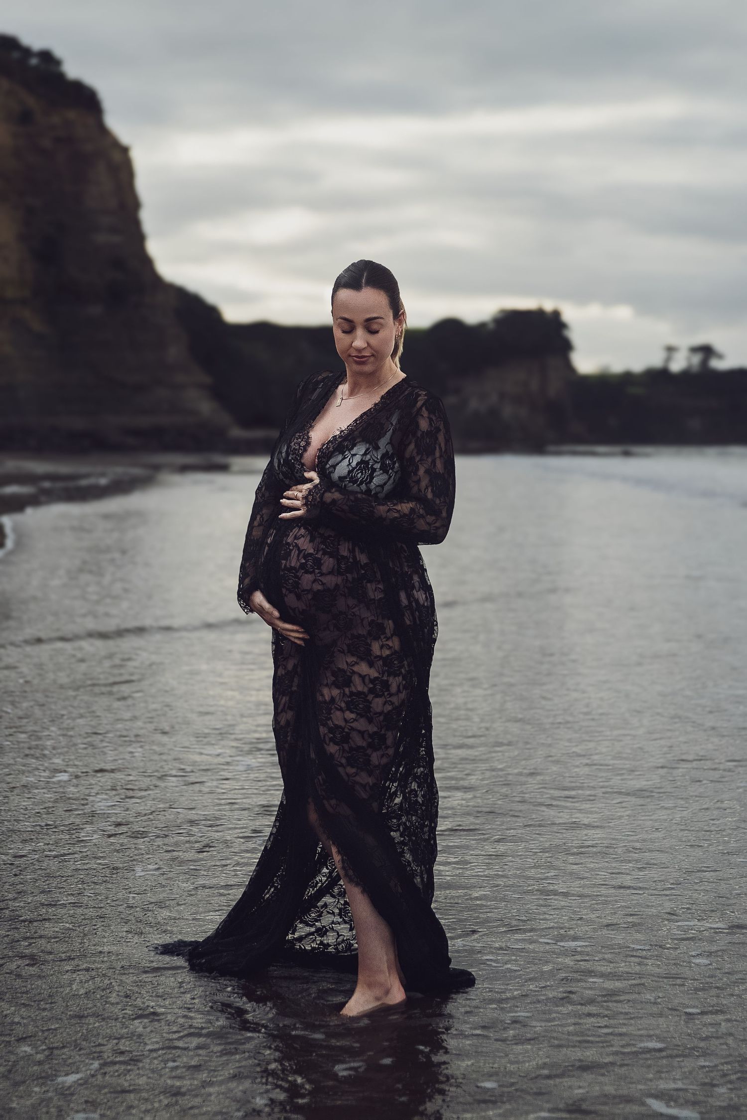 Auckland North Shore Maternity Photoshoot: Pregnant woman in black lace dress standing in the water near dramatic rocks.