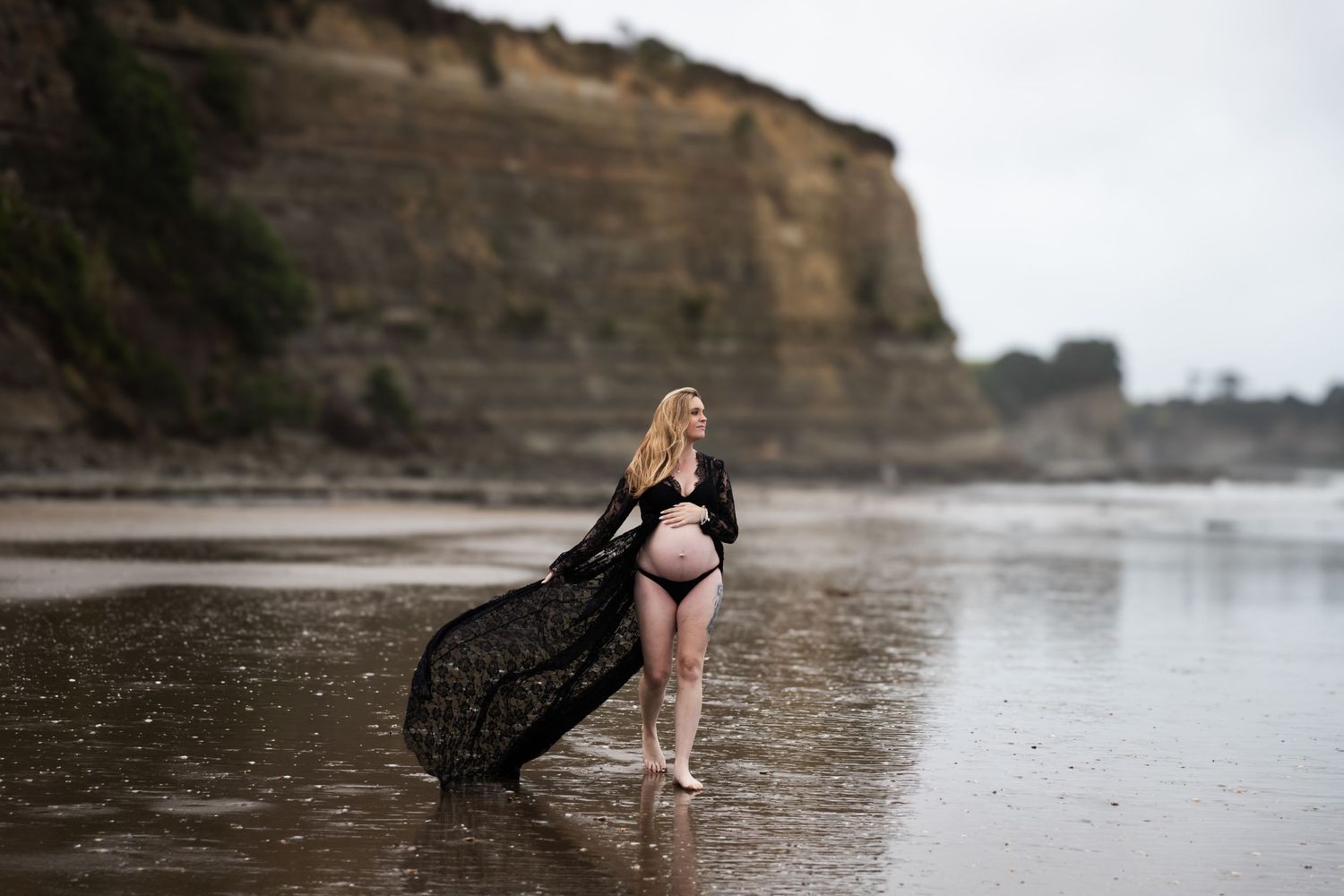 Auckland Maternity Photoshoot: Mum-to-be in black flowing dress on a dramatic North Shore beach, wind lifting the fabric.