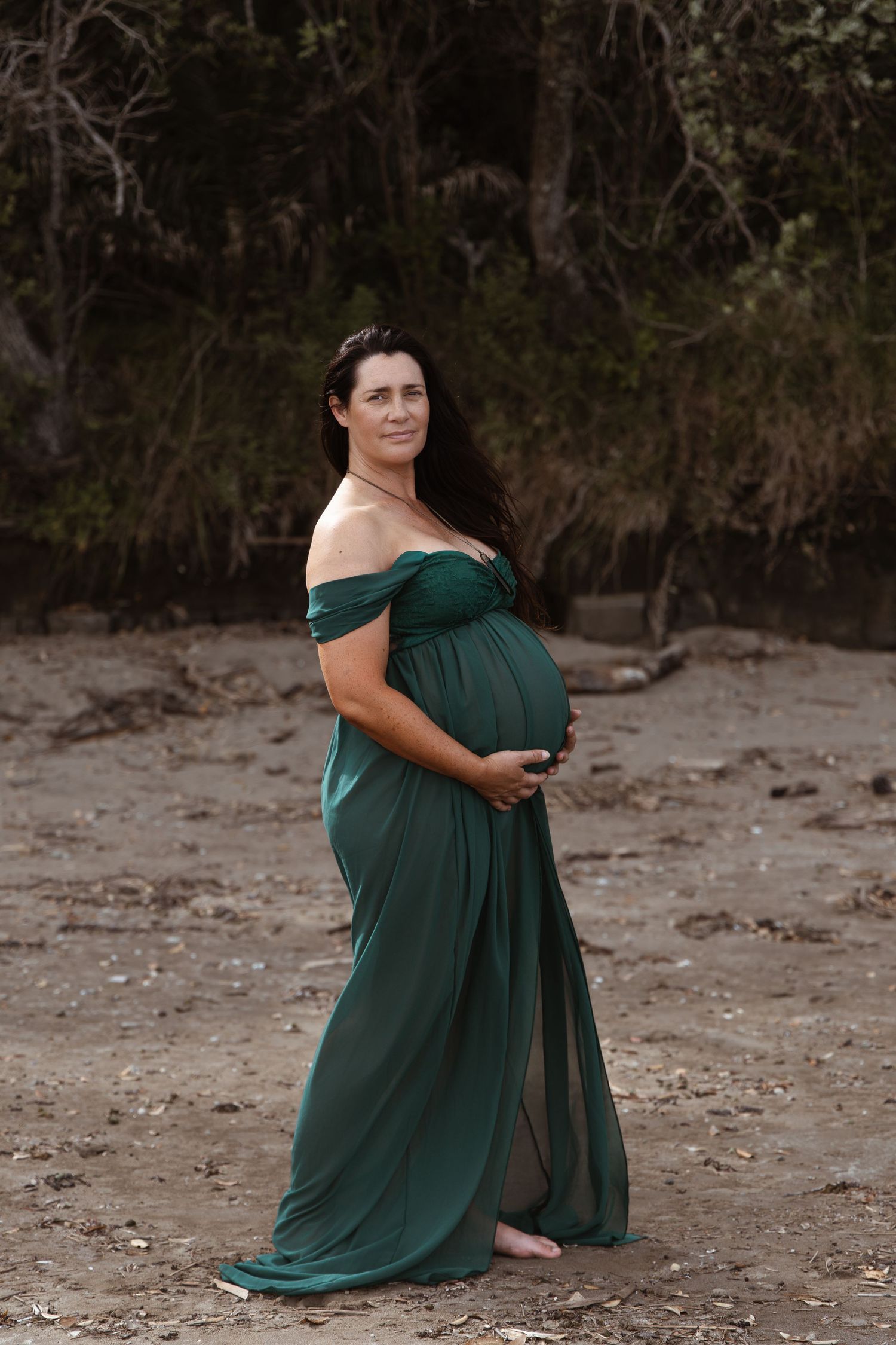 Pregnant woman standing barefoot on a beach in a flowing green dress, photographed by an Auckland photographer